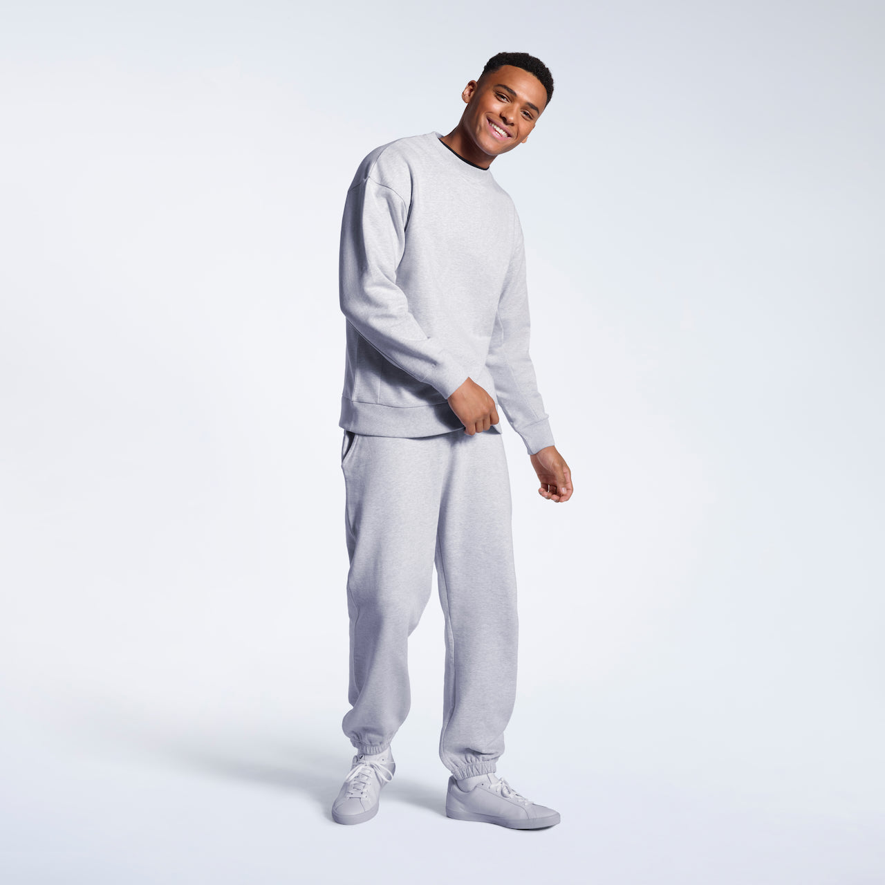 A full body photo of a man smiling into the camera, modelling a grey oversized heavyweight organic cotton sweatshirt with matching joggers.