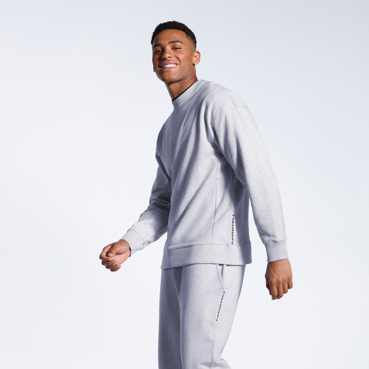 A man smiling into the camera, modelling a grey branded oversized heavyweight organic cotton sweatshirt with matching joggers.