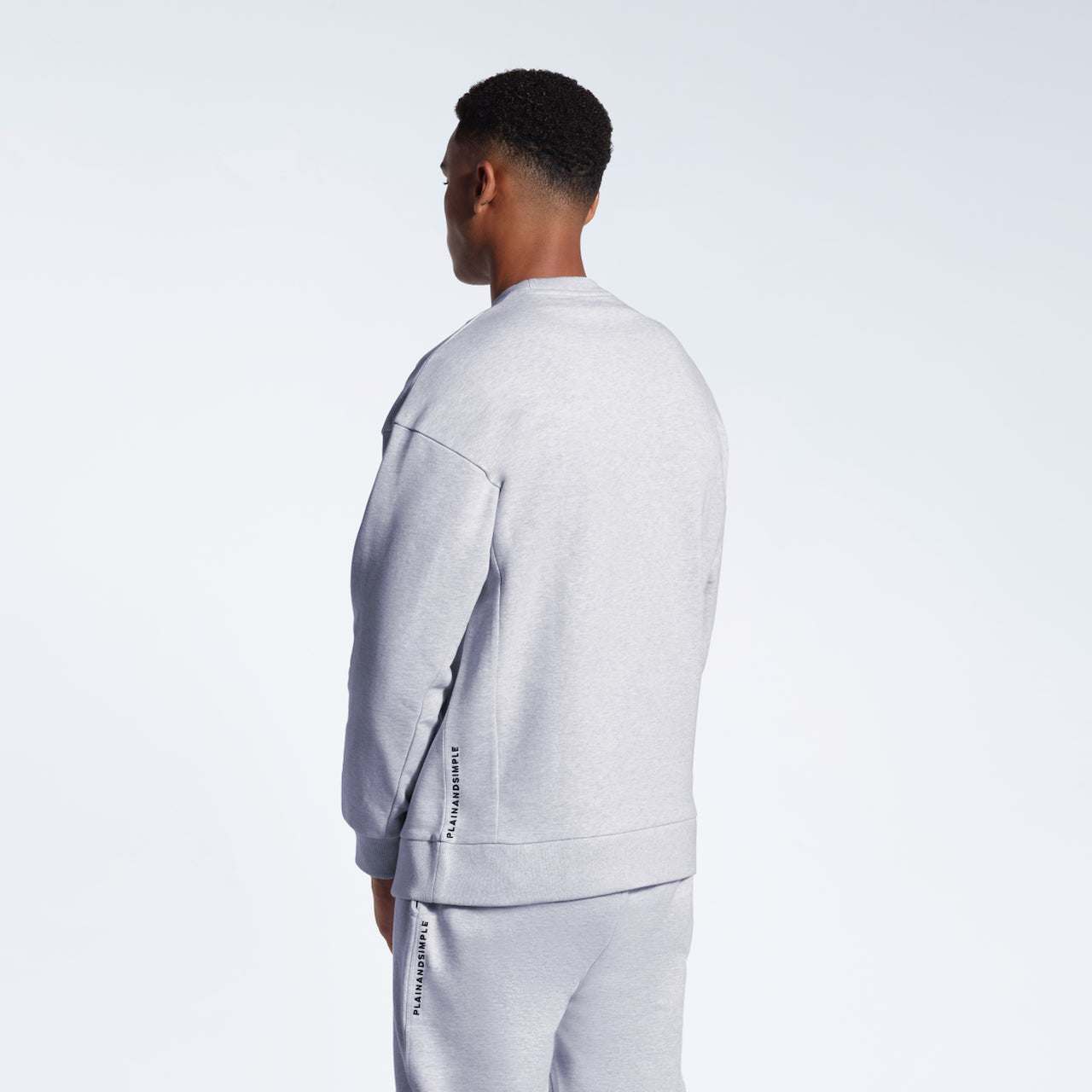 A man facing away from the camera, modelling a grey branded oversized heavyweight organic cotton sweatshirt with matching joggers.