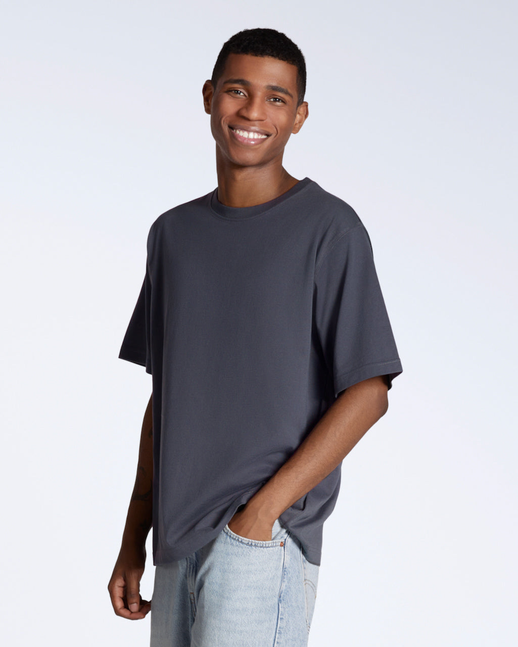 A man with his hand in his pocket smiling into the camera, modelling a plain dark grey, oversized, organic cotton t shirt.