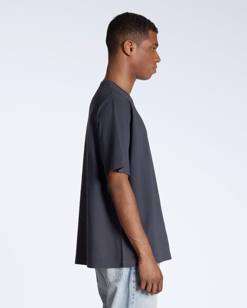 A man facing side on, with his arms by his side, modelling a plain dark grey, oversized, organic cotton t shirt.