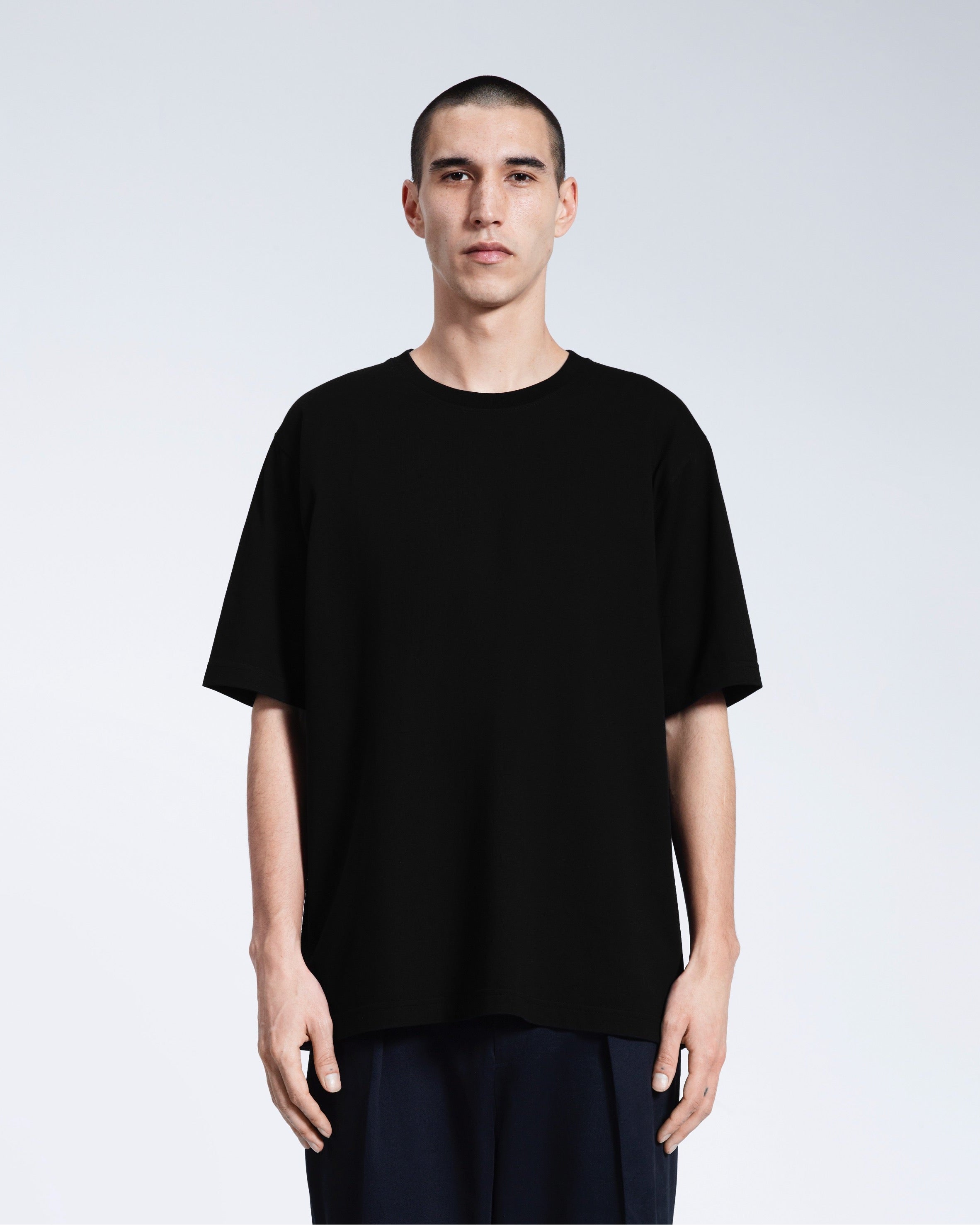A man face on looking into the camera with his arms by his side, modelling a plain black oversized heavyweight organic cotton t shirt.