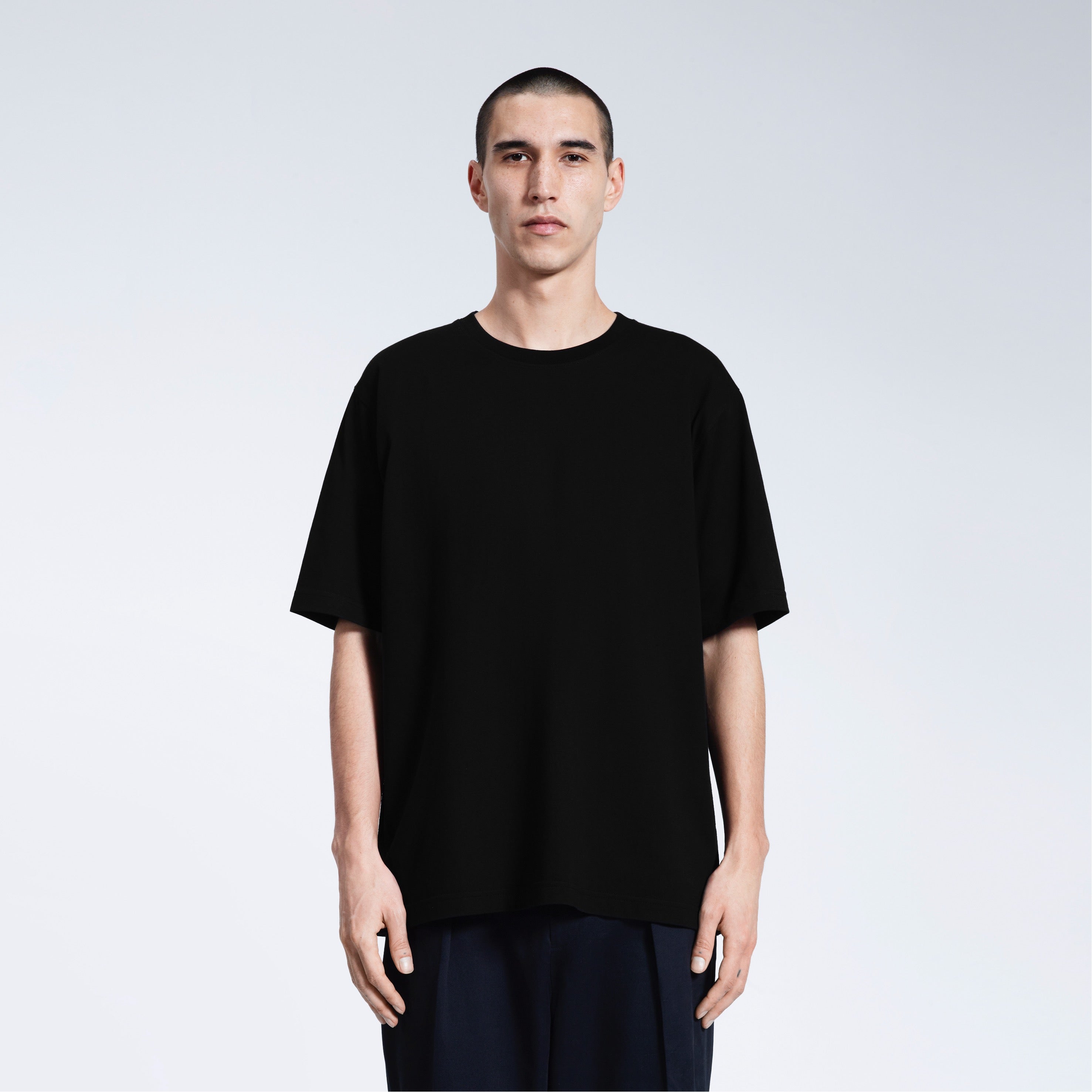 A man face on looking into the camera with his arms by his side, modelling a plain black oversized heavyweight organic cotton t shirt.
