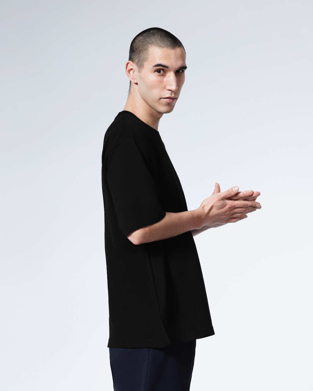 A man facing side on looking at the camera with his hands together, modelling a plain black oversized heavyweight organic cotton t shirt.