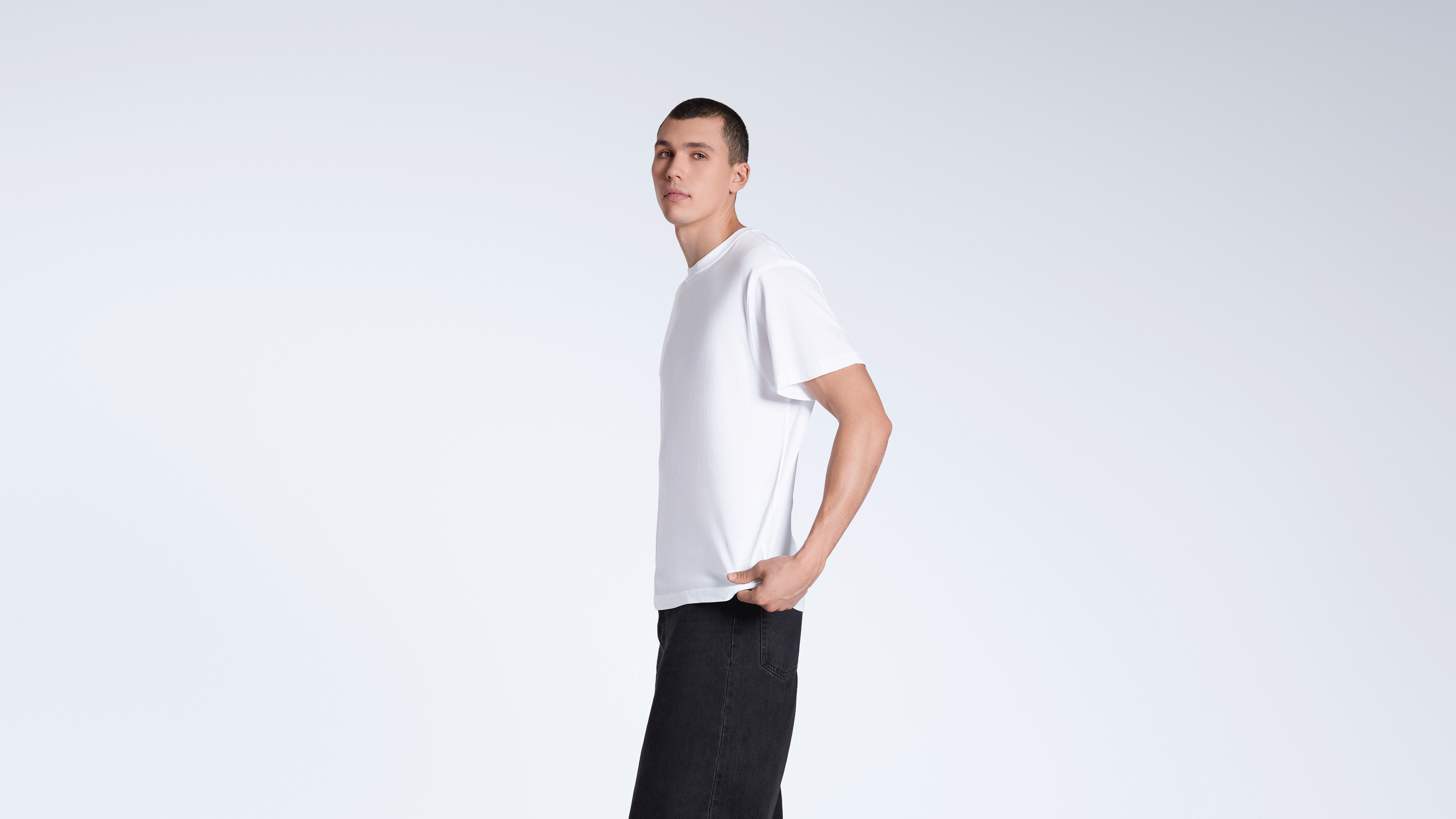 A man facing side on looking into the camera, modelling a plain white, short sleeve organic cotton t shirt.