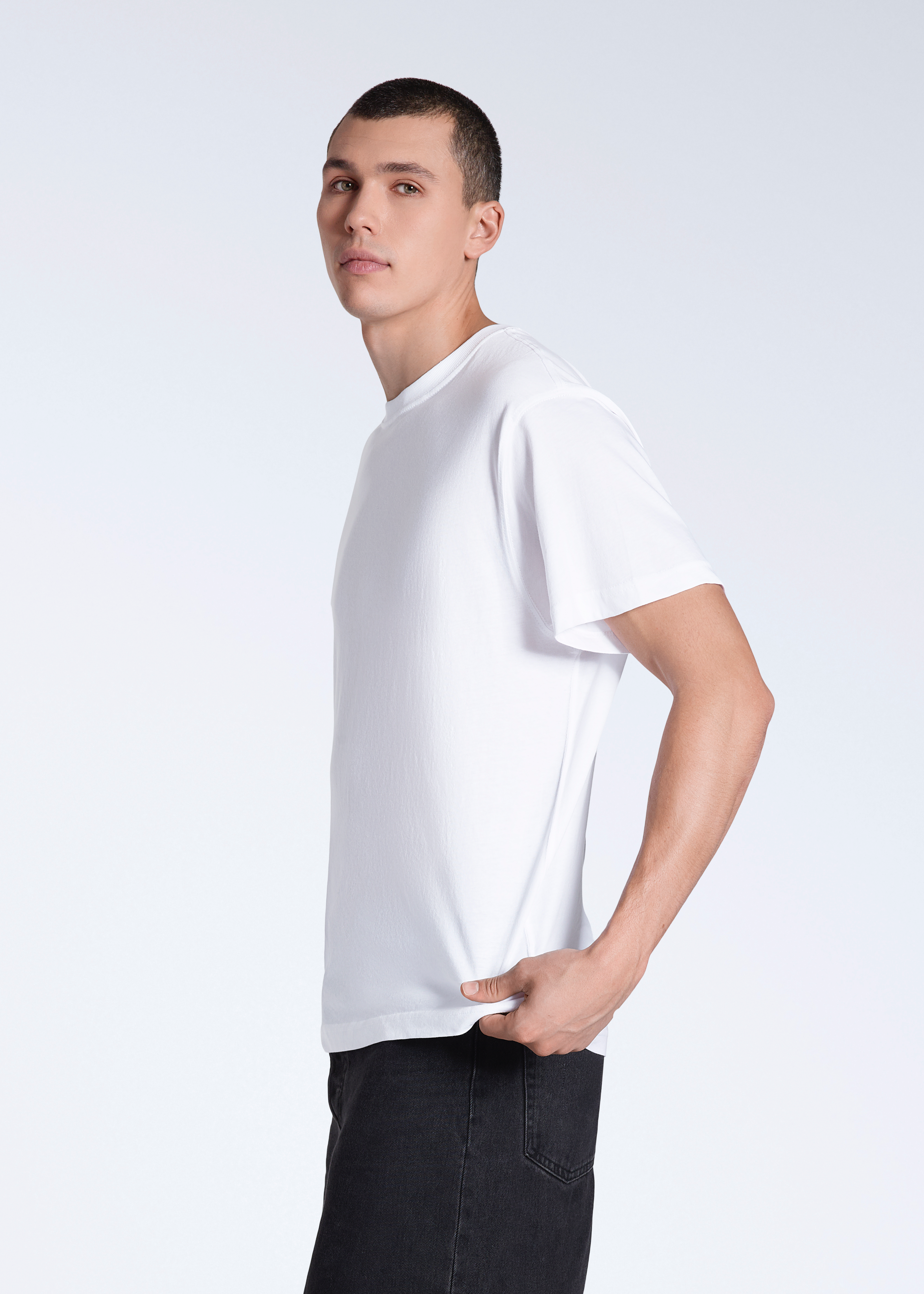 A man facing side on looking into the camera, modelling a plain white, short sleeve organic cotton t shirt.
