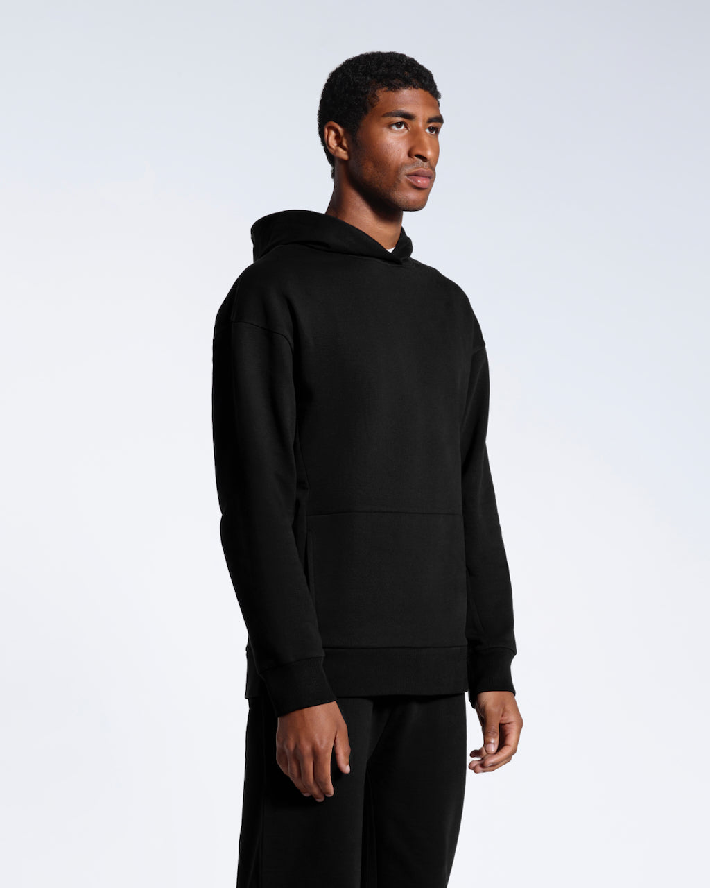 A man at an angle looking away from the camera with his arms by his sides, modelling a black oversized organic cotton hoodie with matching joggers. 