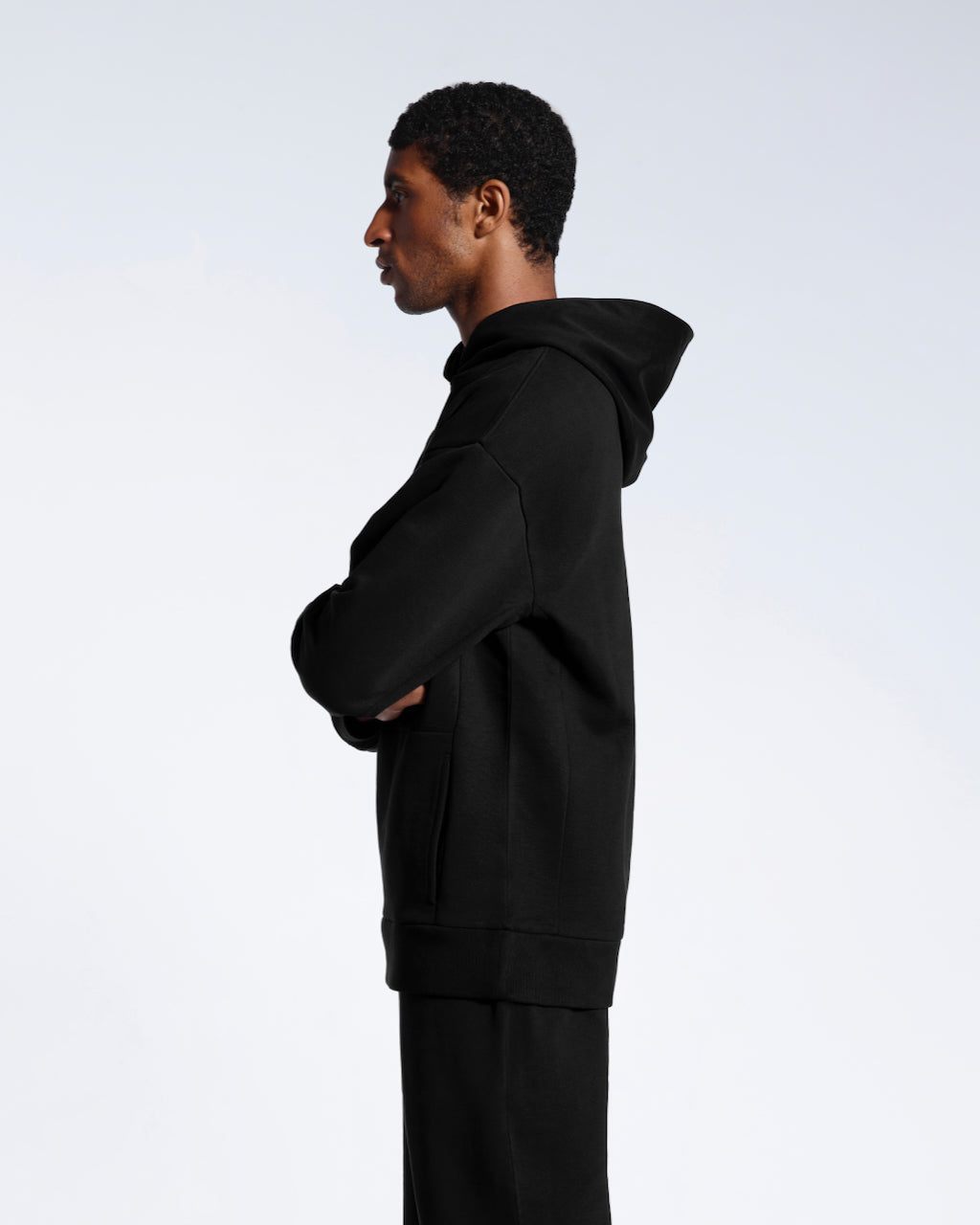 A man facing side on with his arms crossed, modelling a black oversized organic cotton hoodie with matching joggers. 