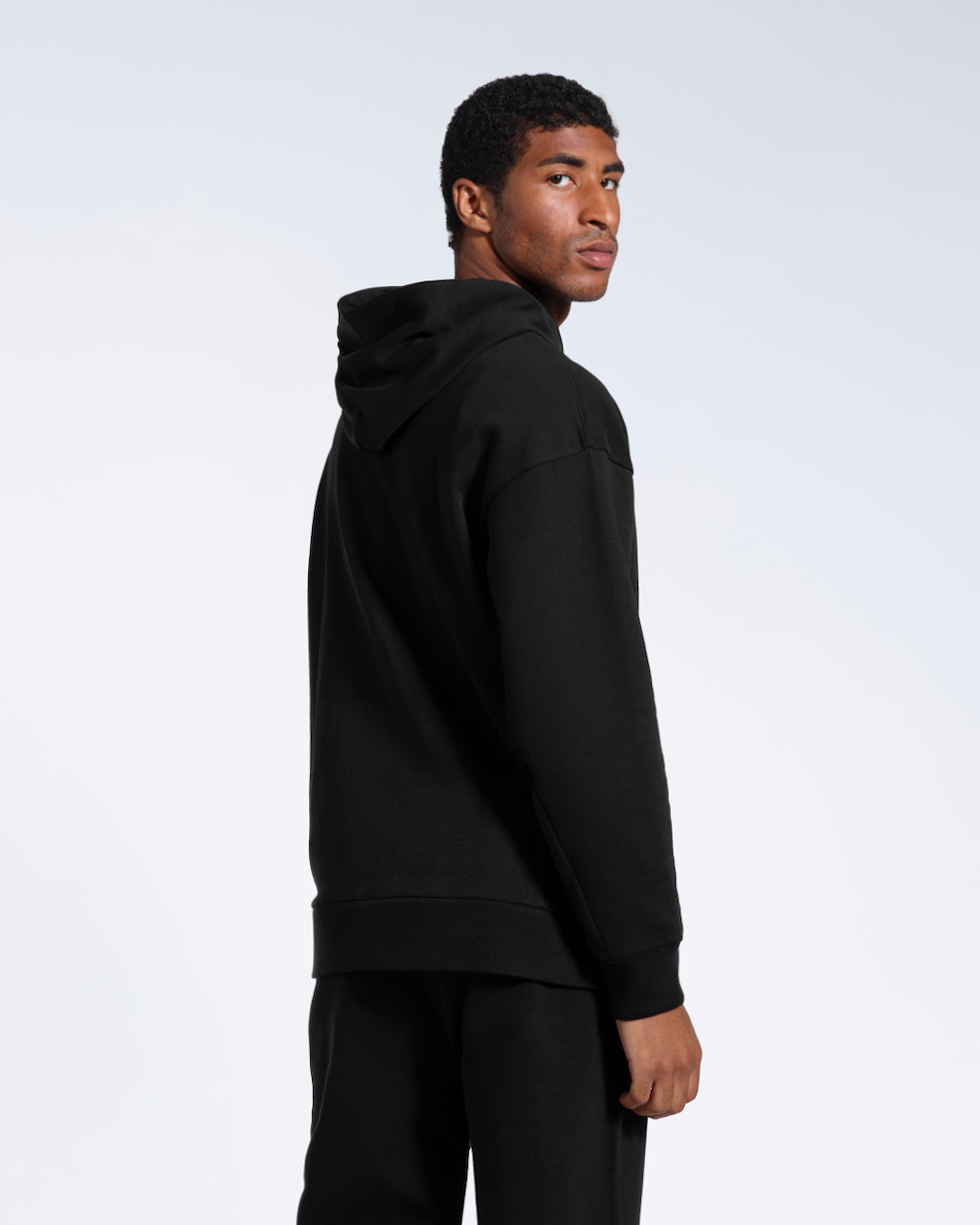 A man looking back at the camera, modelling a black oversized organic cotton hoodie with matching joggers. 