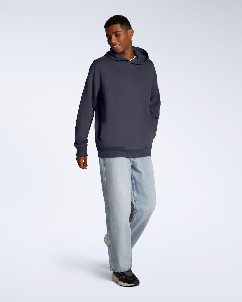 A full body photo of a man walking and smiling, modelling a dark grey oversized organic cotton hoodie with a pair of jeans.