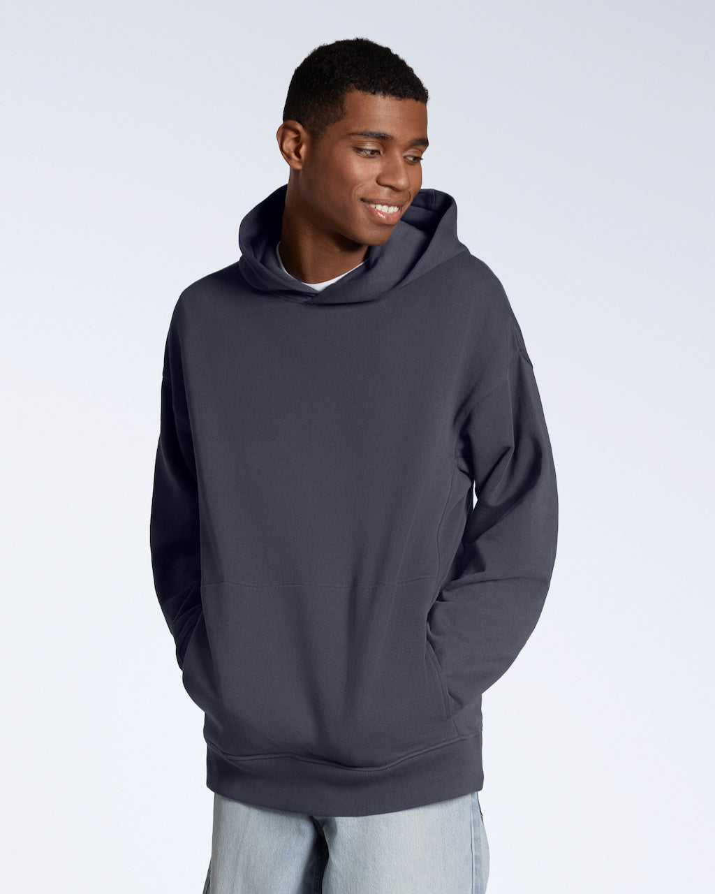 A man smiling with his hands in his pockets, modelling a dark grey oversized organic cotton hoodie.