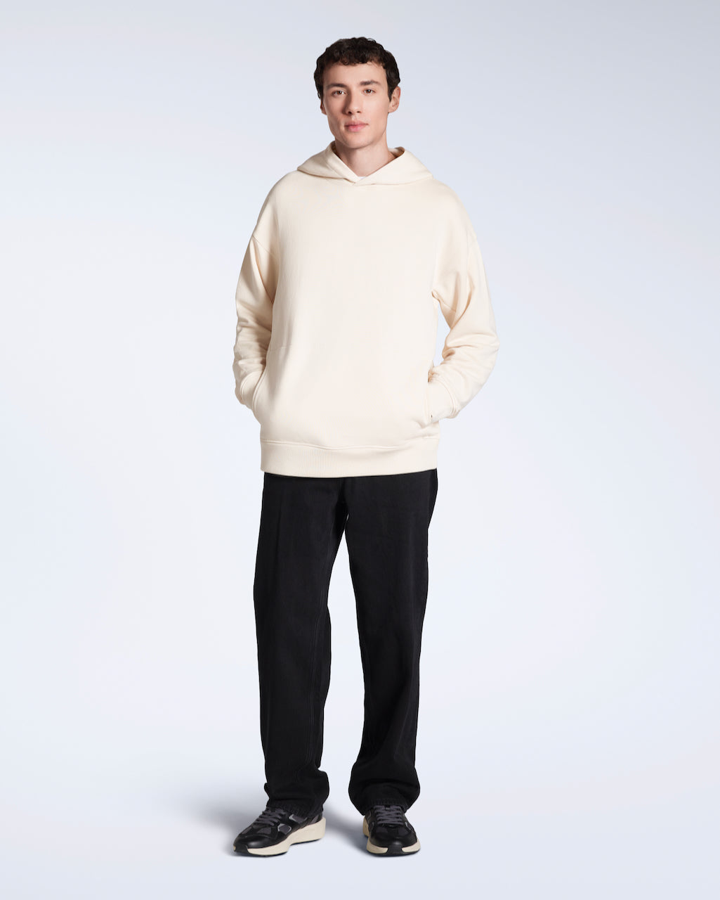 A full body photo of a man with his hands in his pockets looking into the camera, modelling a natural coloured oversized organic cotton hoodie with a pair of jeans.