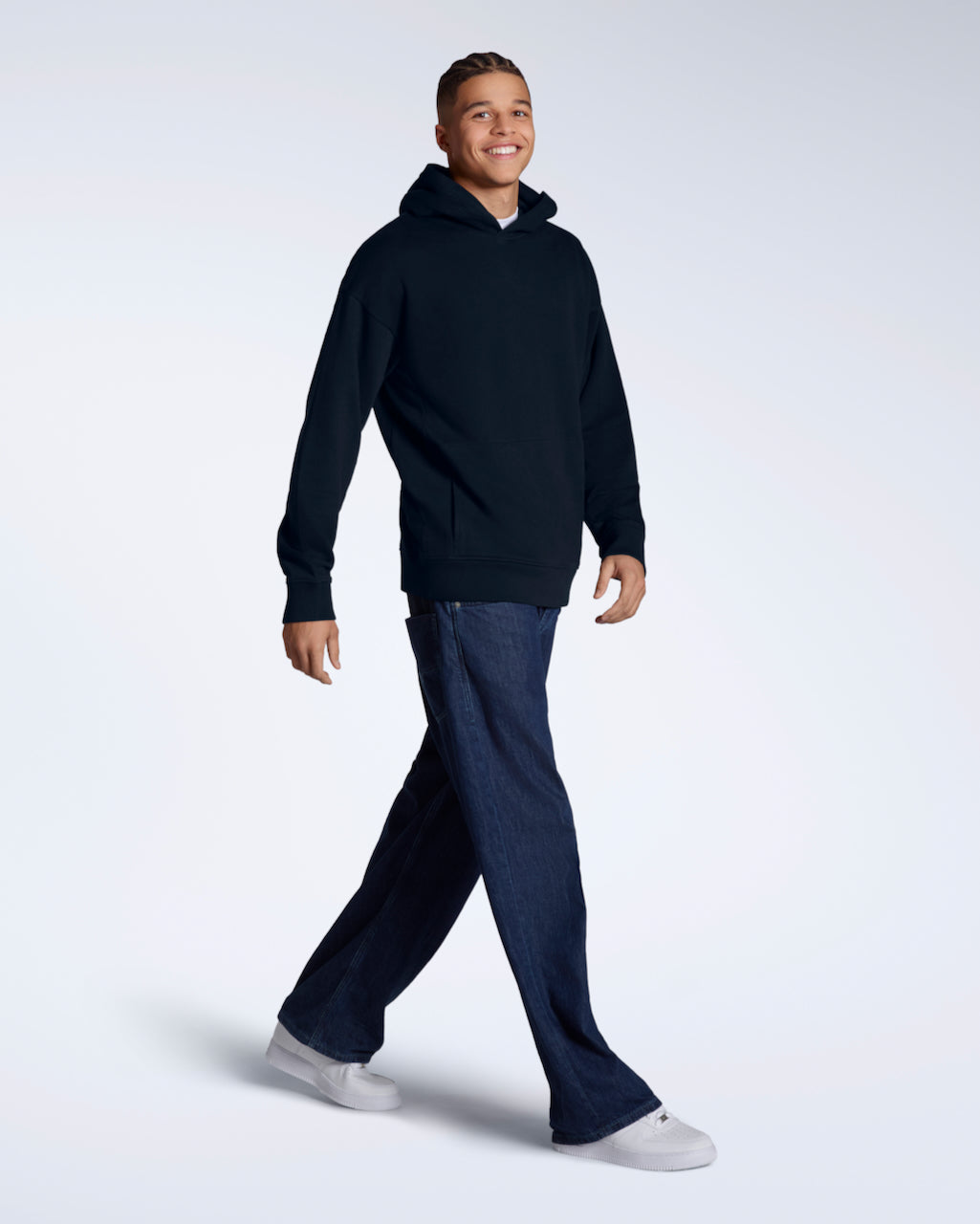 A full body photo of a man walking and smiling into the camera, modelling a navy oversized organic cotton hoodie with a pair of jeans.