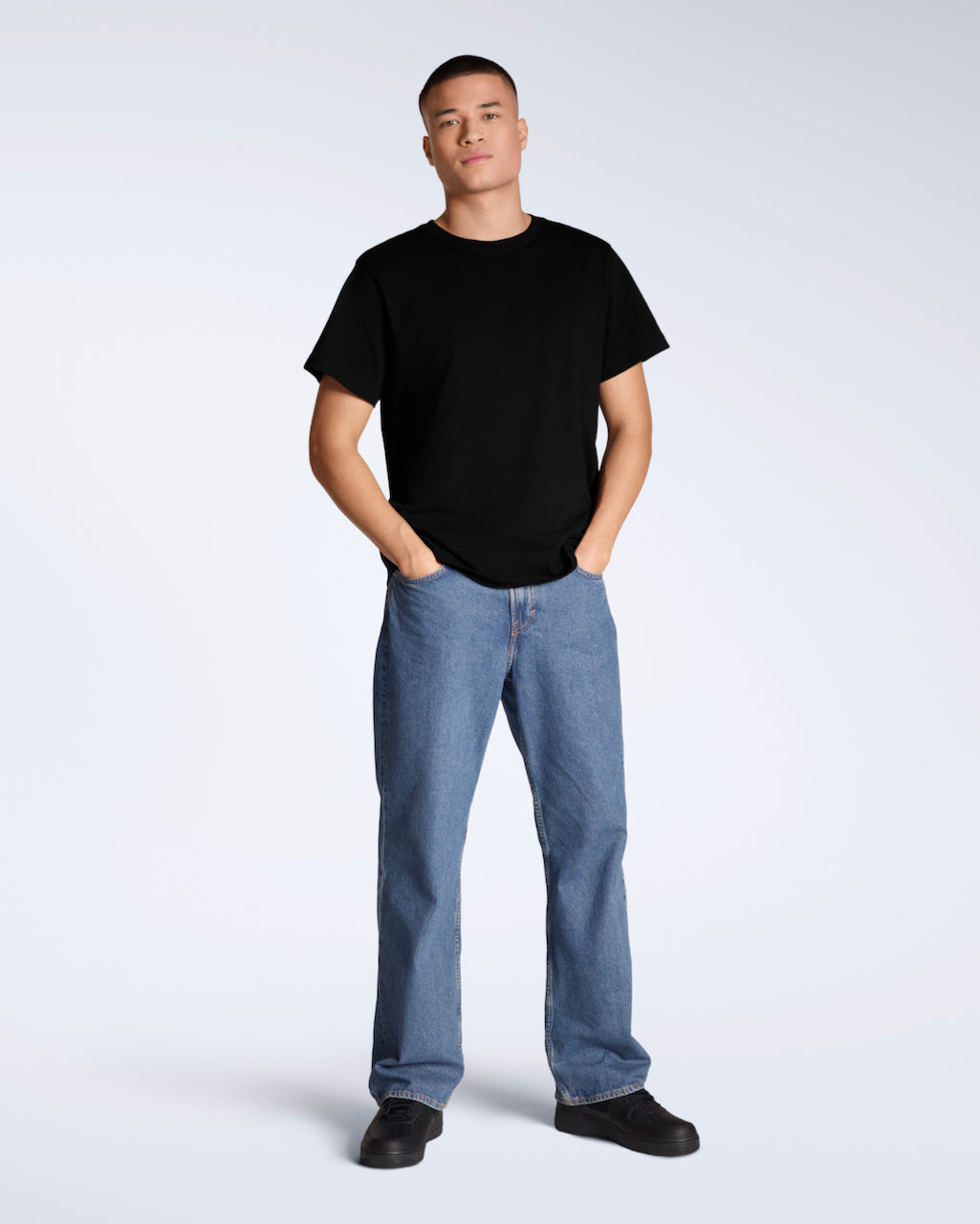 A full body photo of a man looking into the camera with his hands in his pockets, modelling a plain black, short sleeve organic cotton t shirt with a pair of jeans.