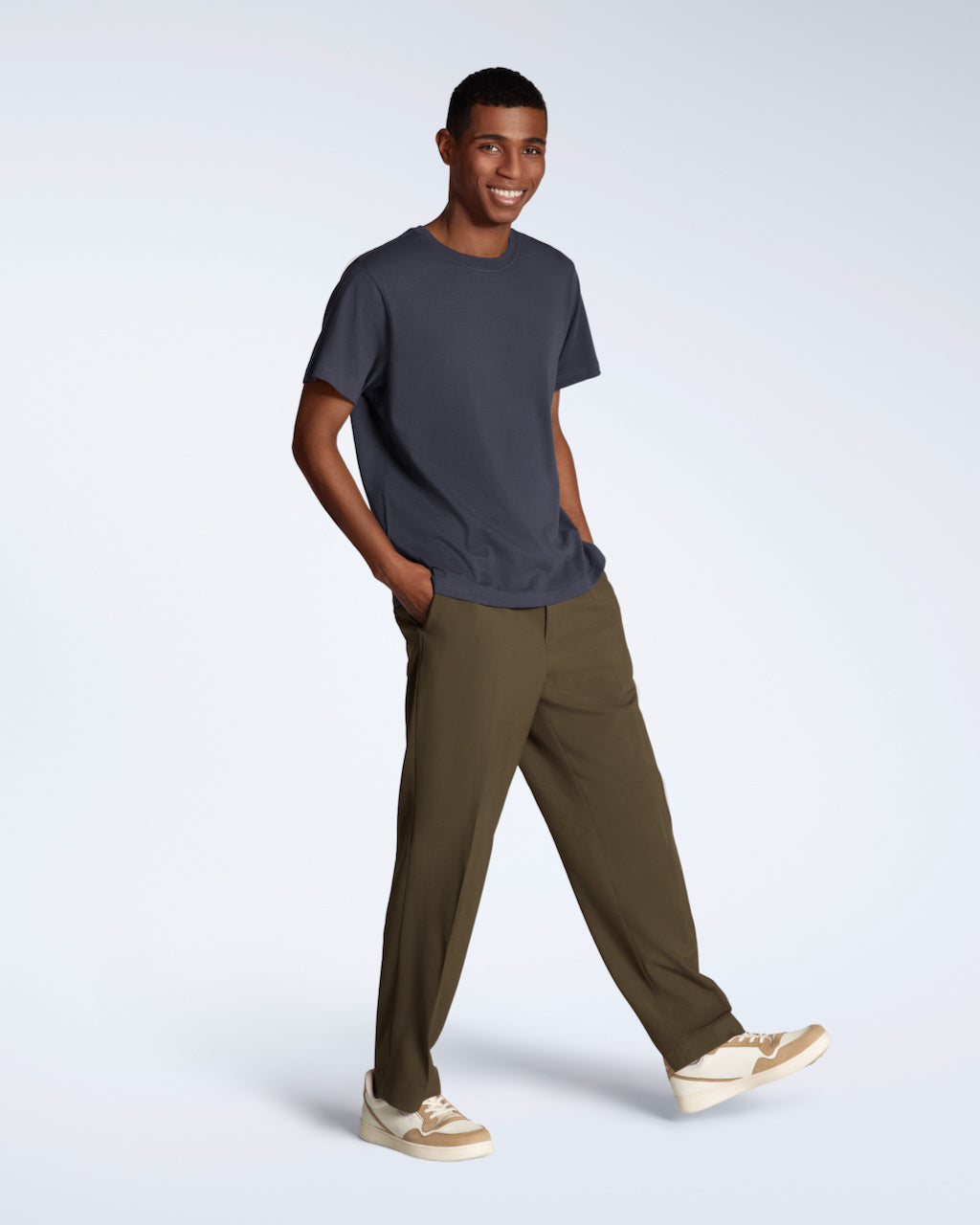 A full body photo of a man smiling into the camera with his hands in his pockets, modelling a plain dark grey, short sleeve organic cotton t shirt with a pair of trousers.