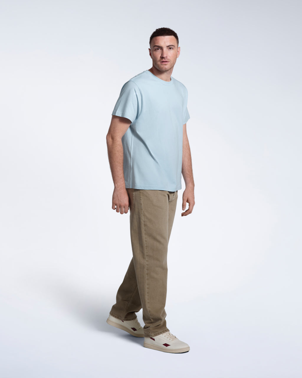 A full body photo of a man looking into the camera, modelling a plain light blue short sleeve organic cotton t shirt.