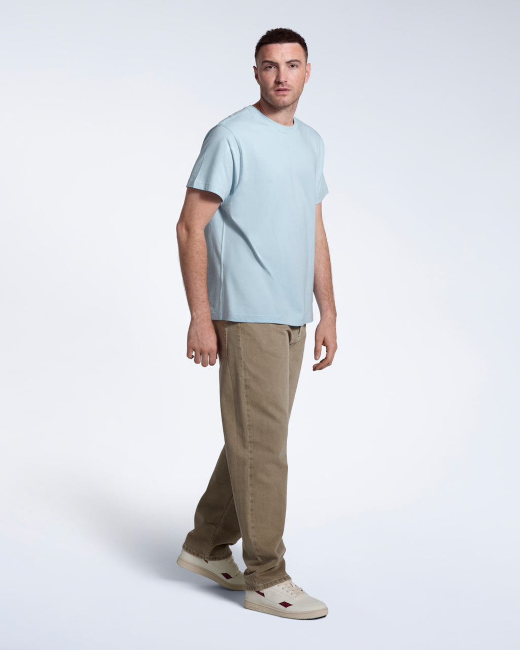 A full body photo of a man looking into the camera, modelling a plain light blue short sleeve organic cotton t shirt.