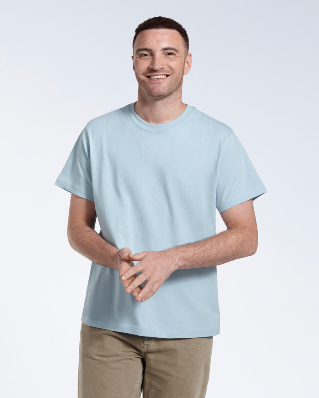 A man smiling into the camera with his hands together, modelling a plain light blue short sleeve organic cotton t shirt.