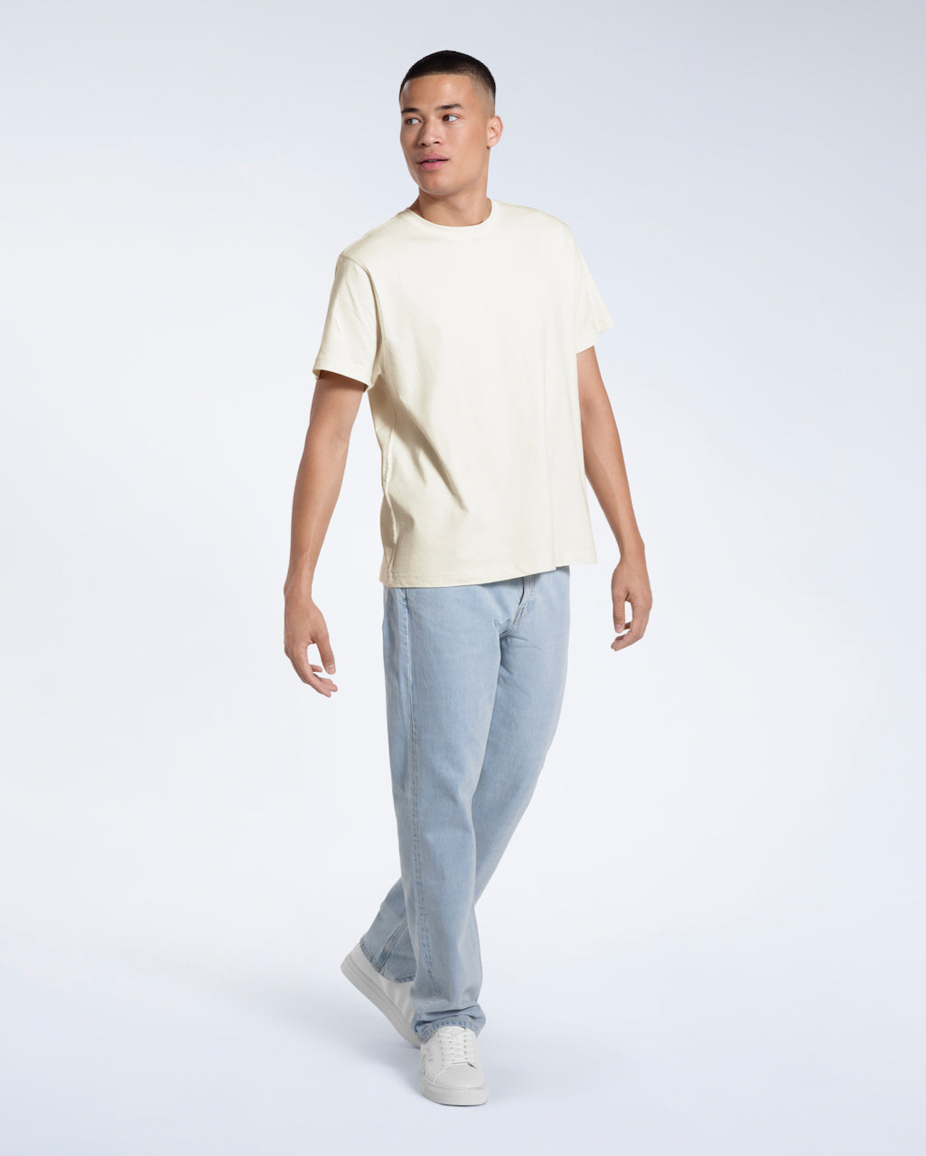 A full body photo of a man walking and looking to the side, modelling a plain ecru short sleeve organic cotton t shirt.