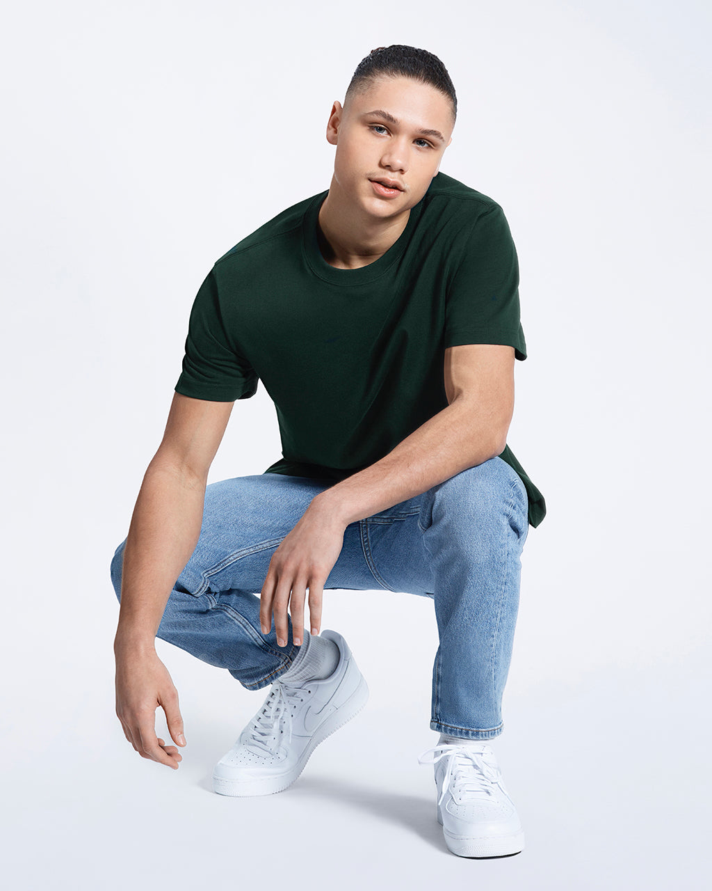 A man crouching down looking into the camera, modelling a plain green short sleeve organic cotton t shirt.