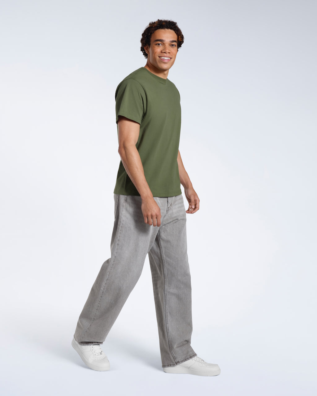 A full body photo of a man looking into the camera smiling, modelling a plain khaki green short sleeve organic cotton t shirt.