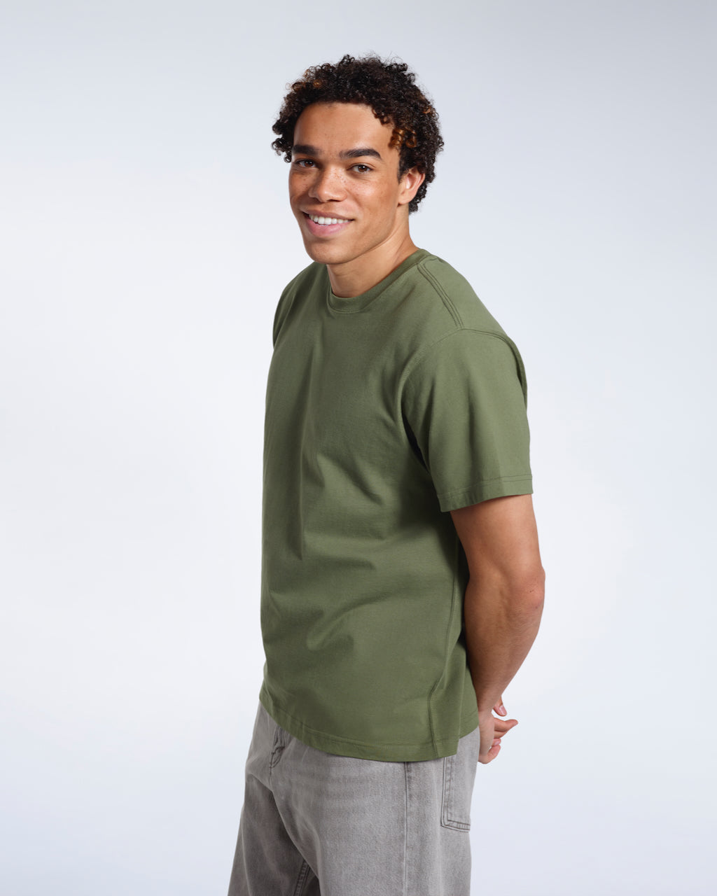 A man smiling into the camera with his hands behind his back, modelling a plain khaki green short sleeve organic cotton t shirt.