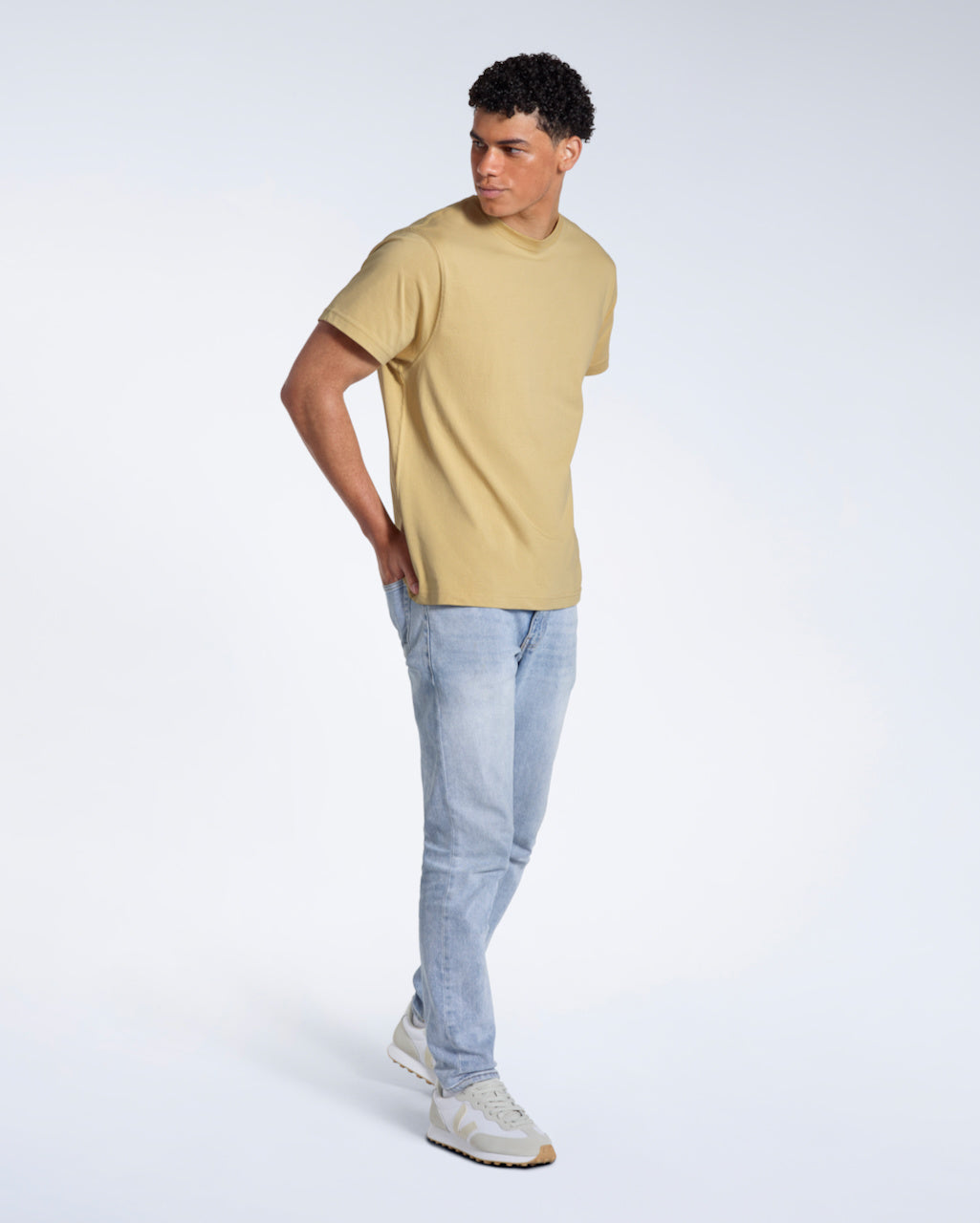 A full body photo of a man looking to the side with his hands in his back pockets, modelling a plain light khaki short sleeve organic cotton t shirt.