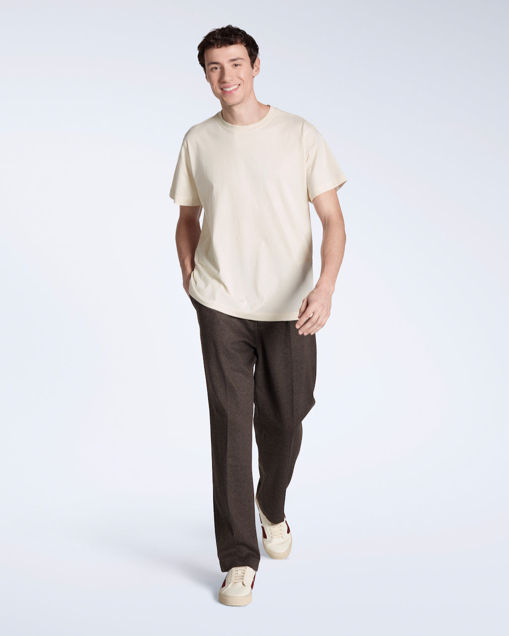 A full body photo of a man smiling into the camera with his hand in his pocket, modelling a plain natural, short sleeve organic cotton t shirt with a pair of trousers.