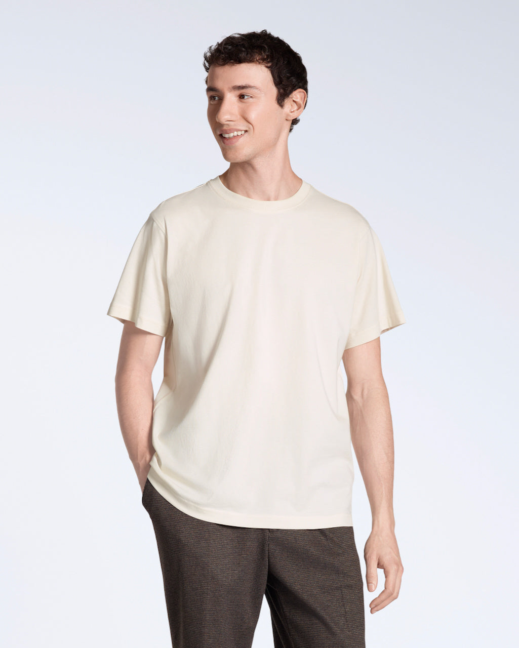 A man smiling with his hand in his pocket, modelling a plain natural coloured, short sleeve organic cotton t shirt.