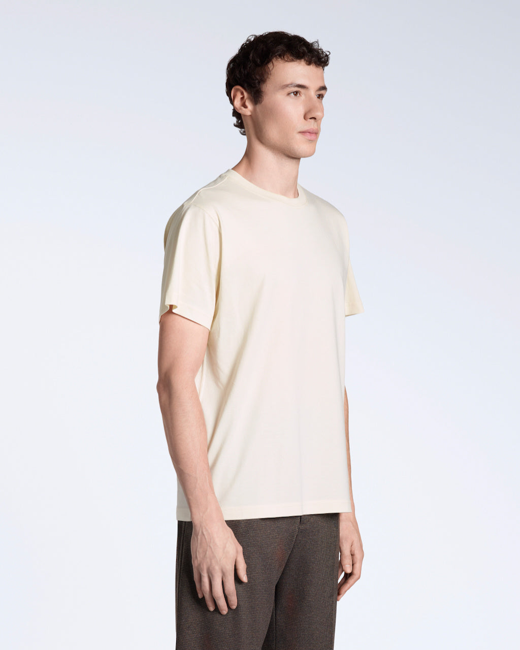A man at an angle with his arms by his sides, modelling a plain natural coloured, short sleeve organic cotton t shirt.