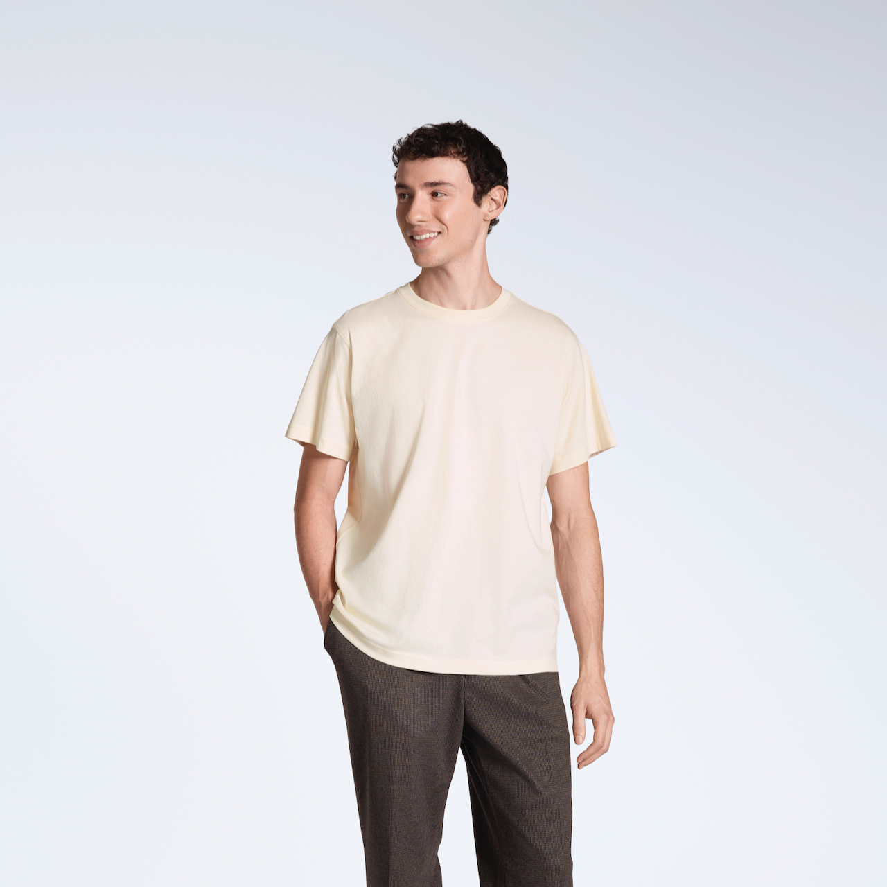 A man smiling with his hand in his pocket, modelling a plain natural coloured, short sleeve organic cotton t shirt.