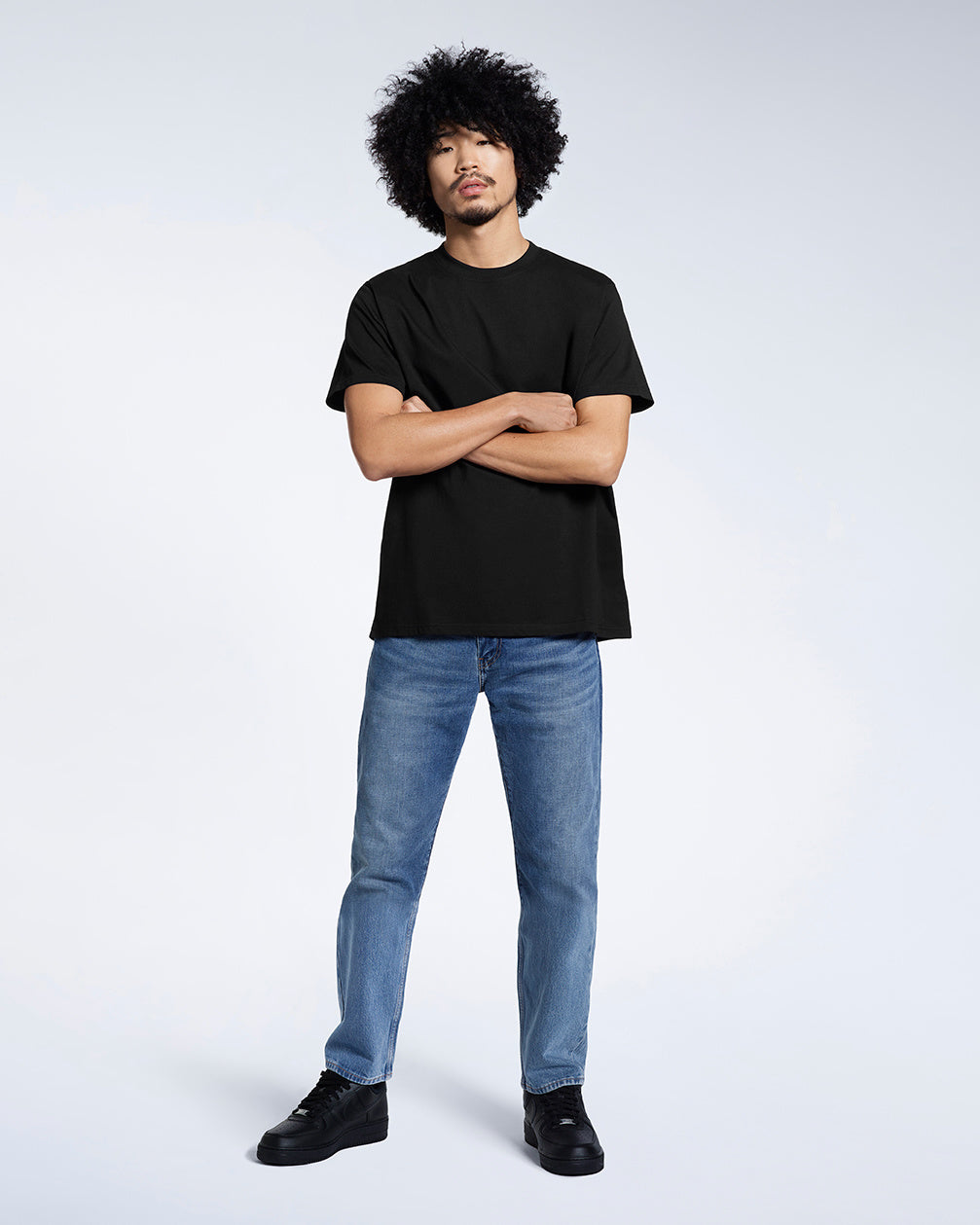 A full body photo of a man looking into the camera with his arms crossed, modelling a plain black short sleeve organic cotton t shirt.