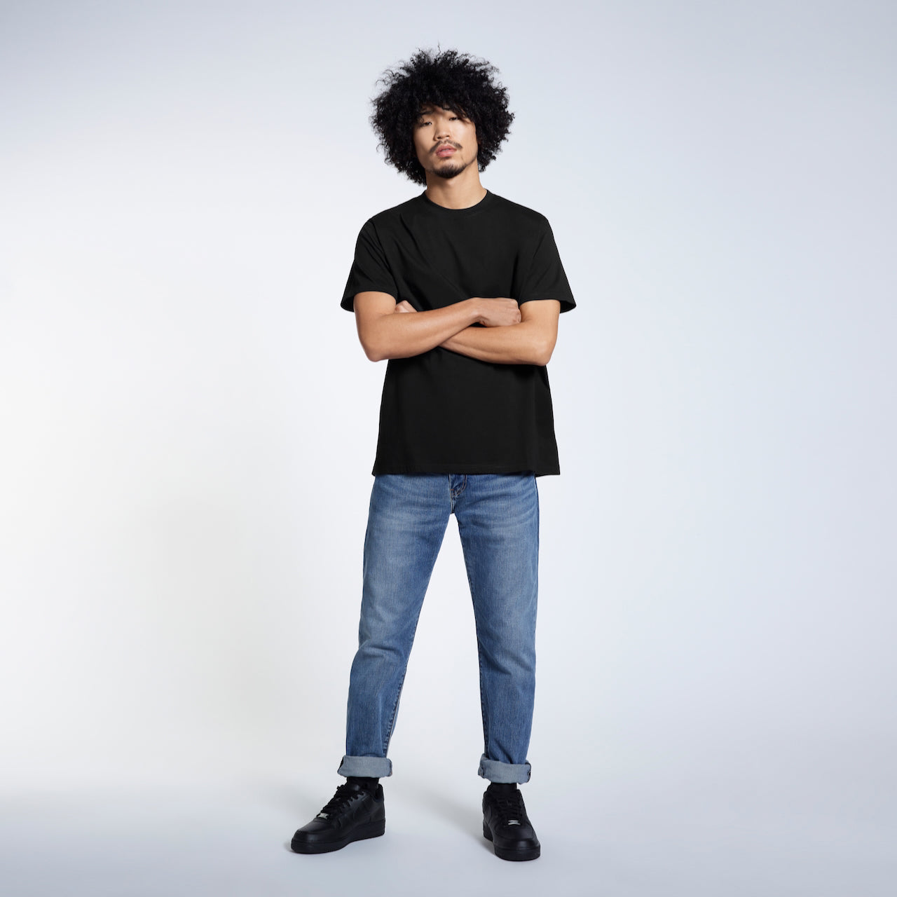 A full body photo of a man looking into the camera with his arms crossed, modelling a plain black short sleeve organic cotton t shirt.