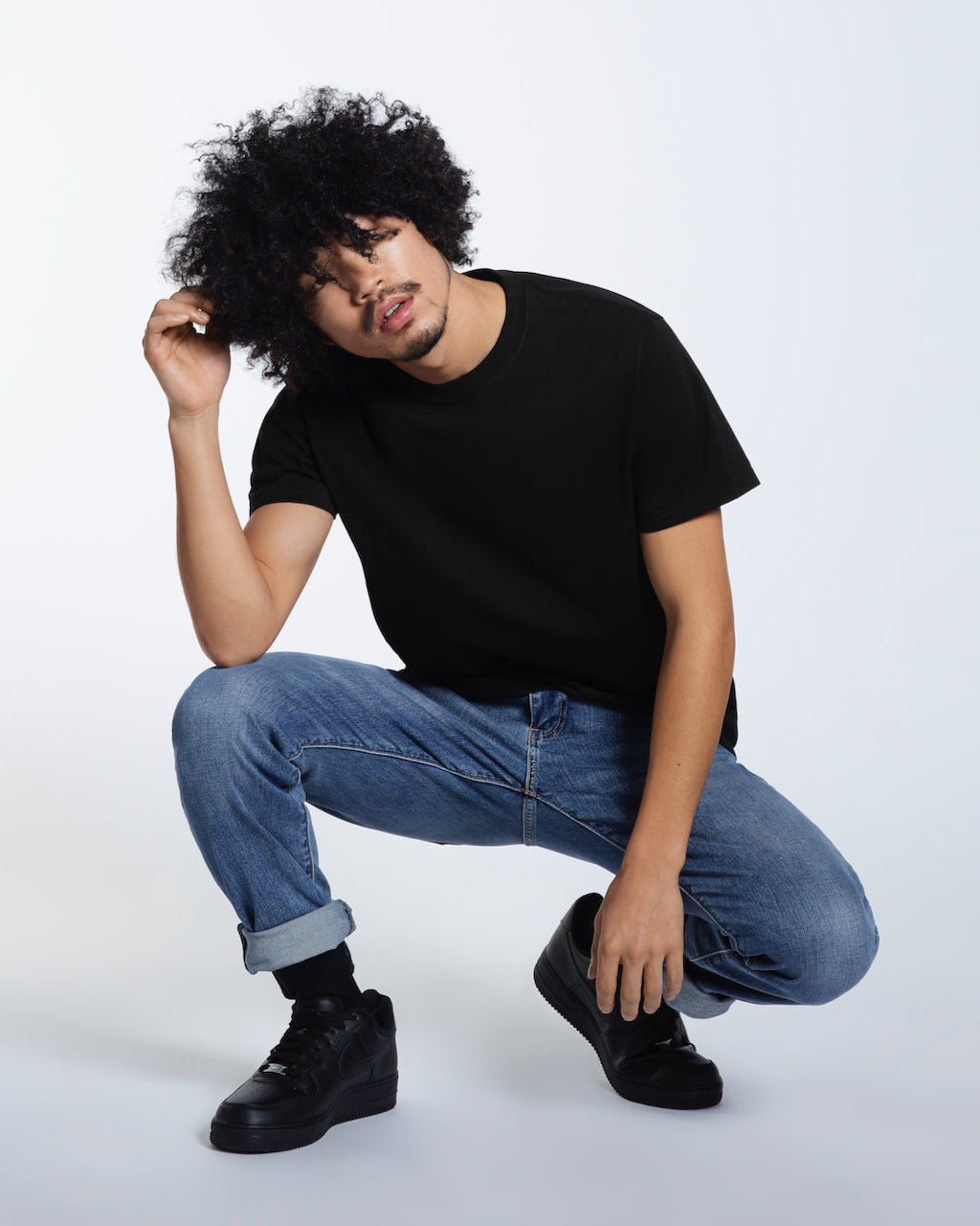 A man crouching down and touching his hair, modelling a plain black short sleeve organic cotton t shirt.