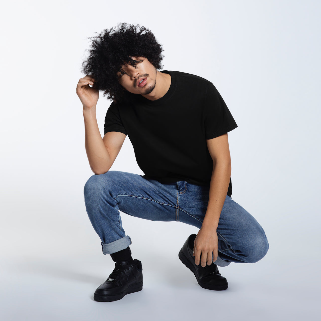 A man crouching down and touching his hair, modelling a plain black short sleeve organic cotton t shirt.