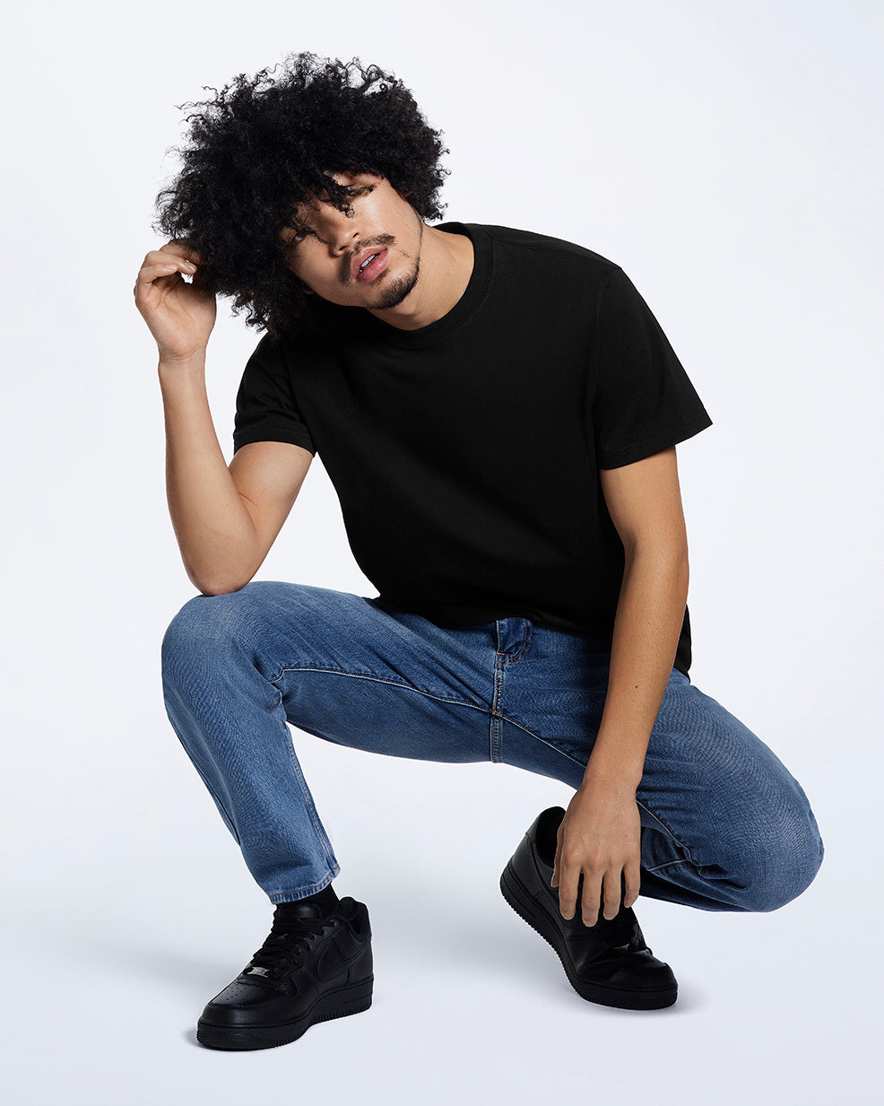 A man crouching down and touching his hair, modelling a plain black short sleeve organic cotton t shirt.