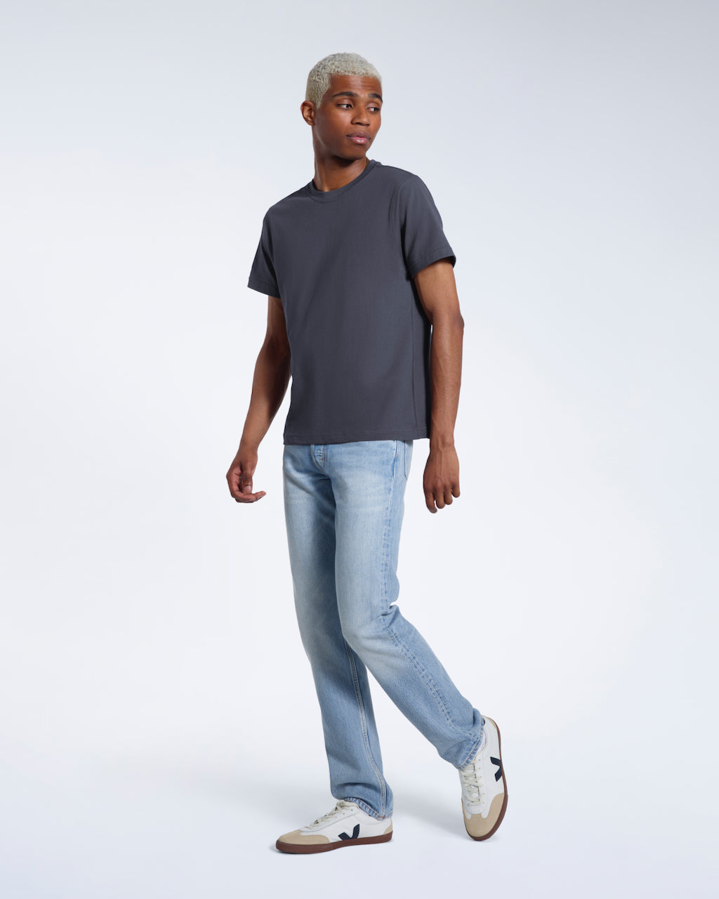 A full body photo of a man looking to the side, modelling a plain dark grey short sleeve organic cotton t shirt.