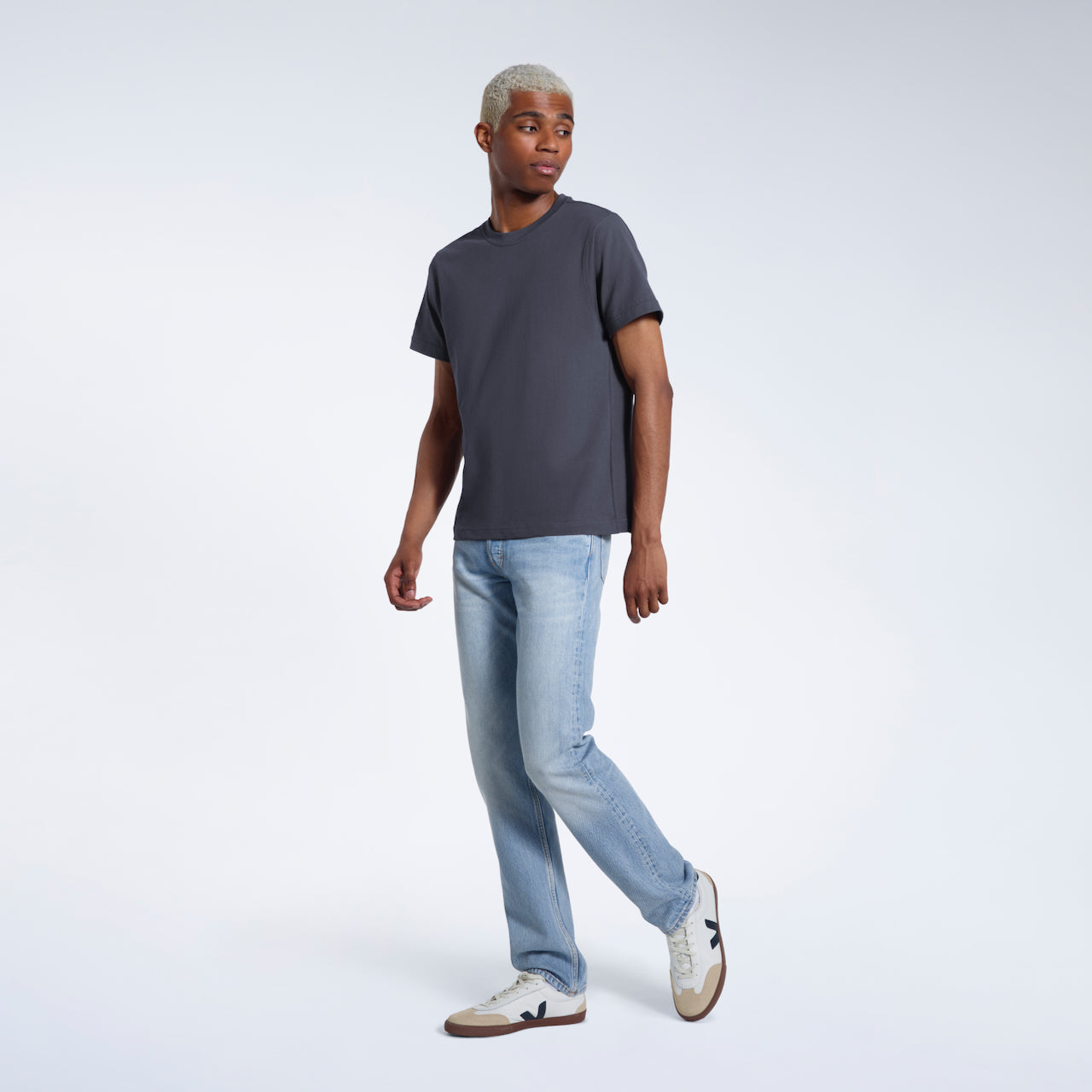 A full body photo of a man looking to the side, modelling a plain dark grey short sleeve organic cotton t shirt.