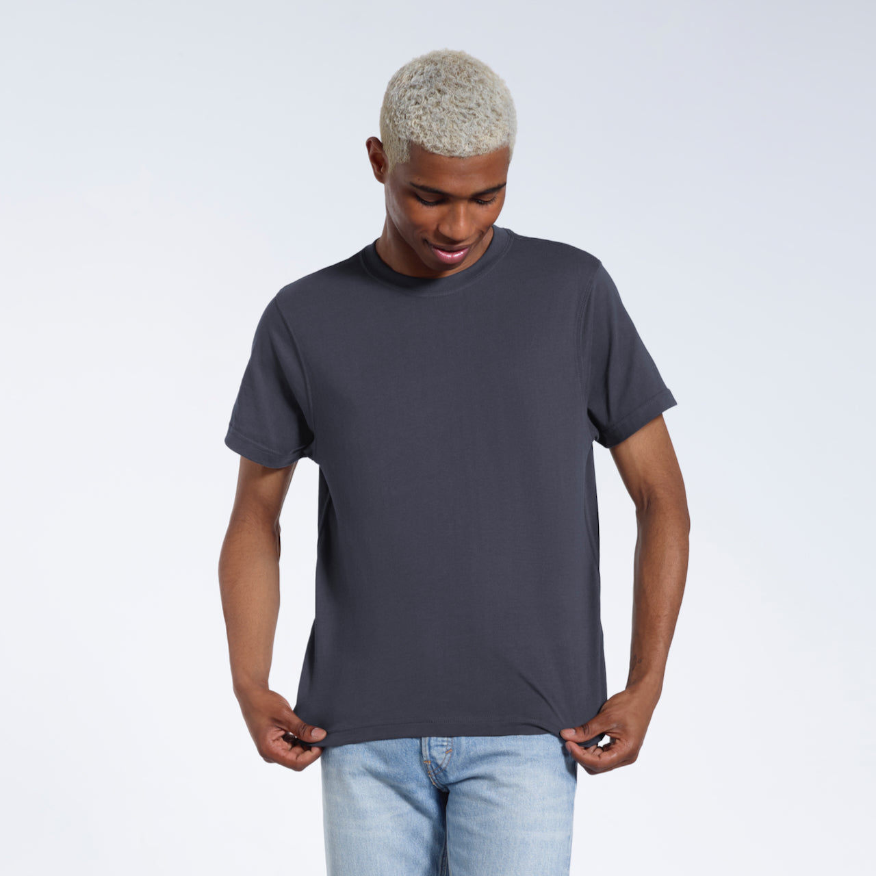 A man looking down and touching the bottom of his plain dark grey short sleeve organic cotton t shirt.