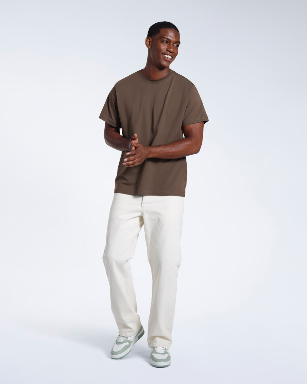 A full body photo of a man smiling with his hands together, modelling a plain dark khaki short sleeve organic cotton t shirt.