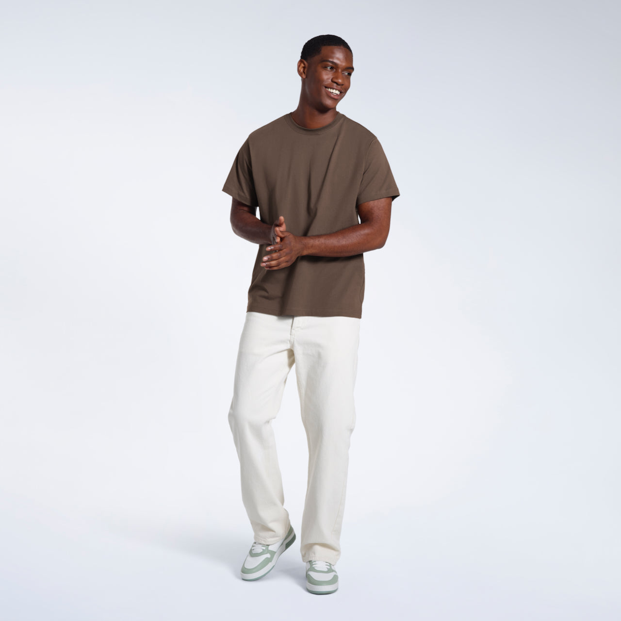 A full body photo of a man smiling with his hands together, modelling a plain dark khaki short sleeve organic cotton t shirt.