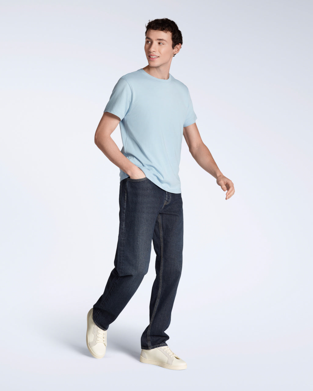 A full body photo of a man smiling with his hand in his pocket, modelling a plain light blue, short sleeve organic cotton t shirt with a pair of jeans.