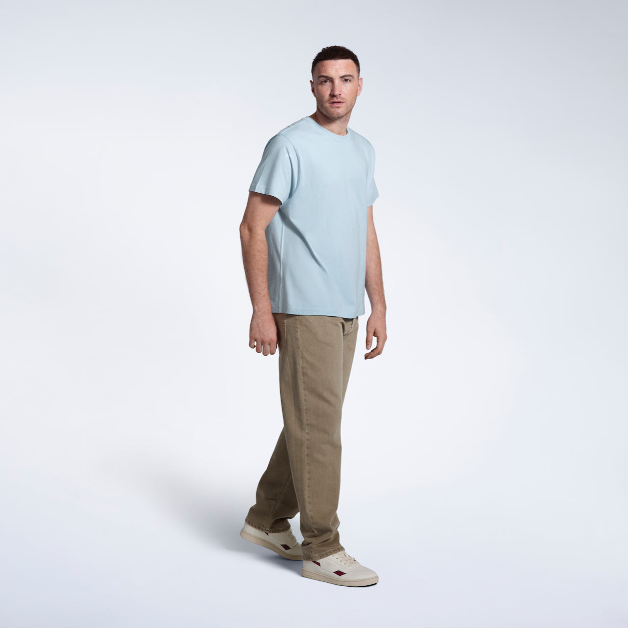 A full body photo of a man looking into the camera, modelling a plain light blue short sleeve organic cotton t shirt.