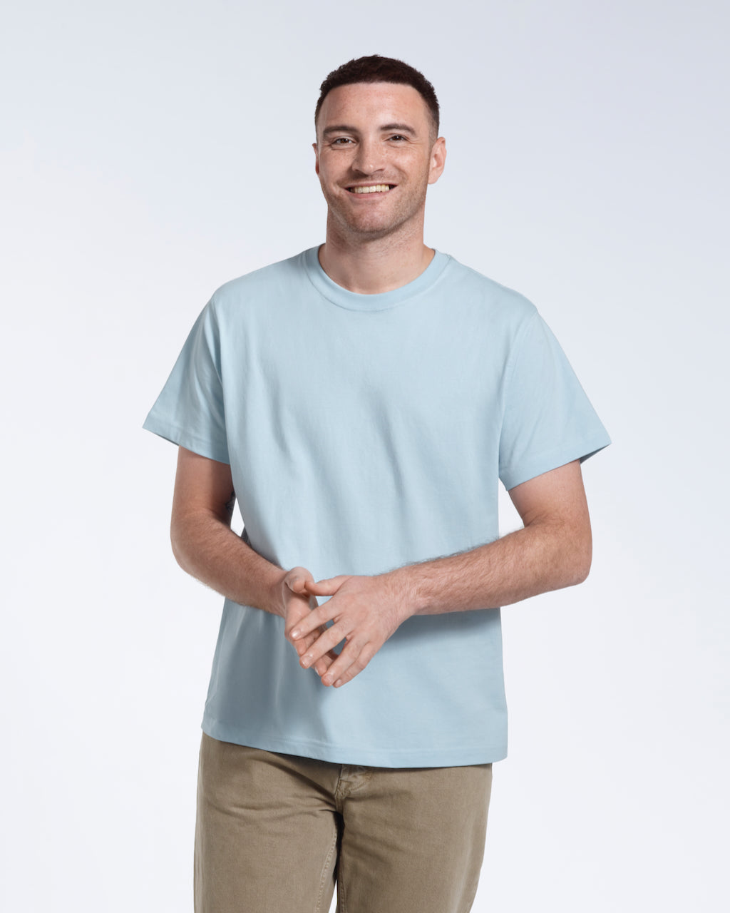 A man smiling into the camera with his hands together, modelling a plain light blue short sleeve organic cotton t shirt.