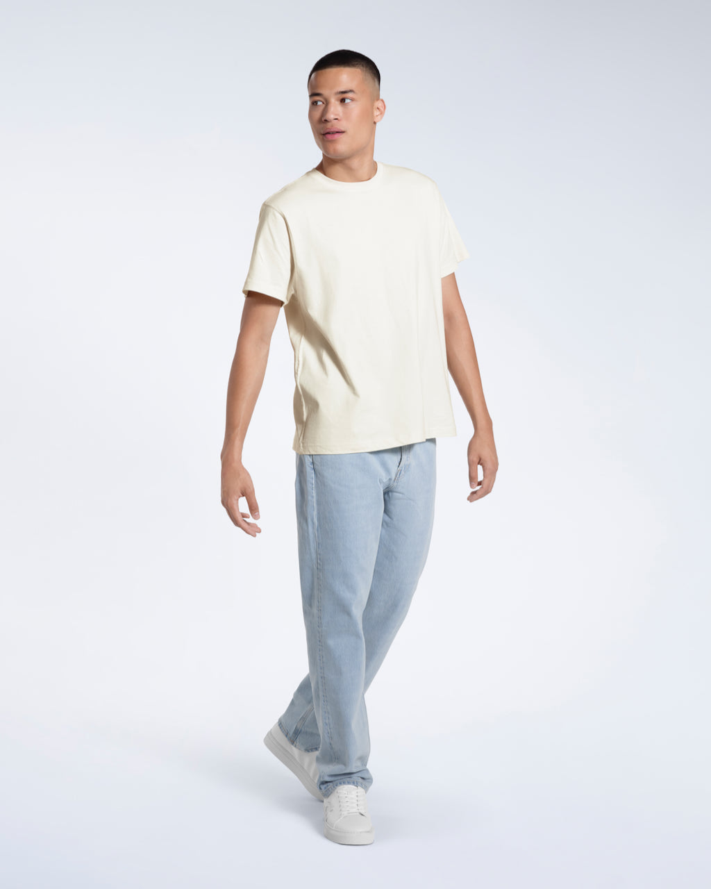 A full body photo of a man walking and looking to the side, modelling a plain ecru short sleeve organic cotton t shirt.