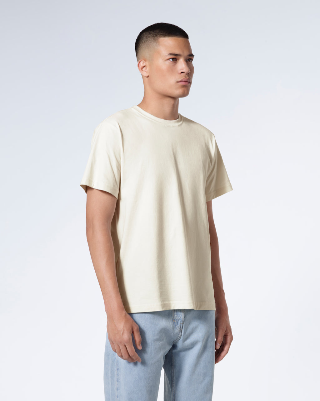 A man at an angle with his arms by his sides, modelling a plain ecru short sleeve organic cotton t shirt.