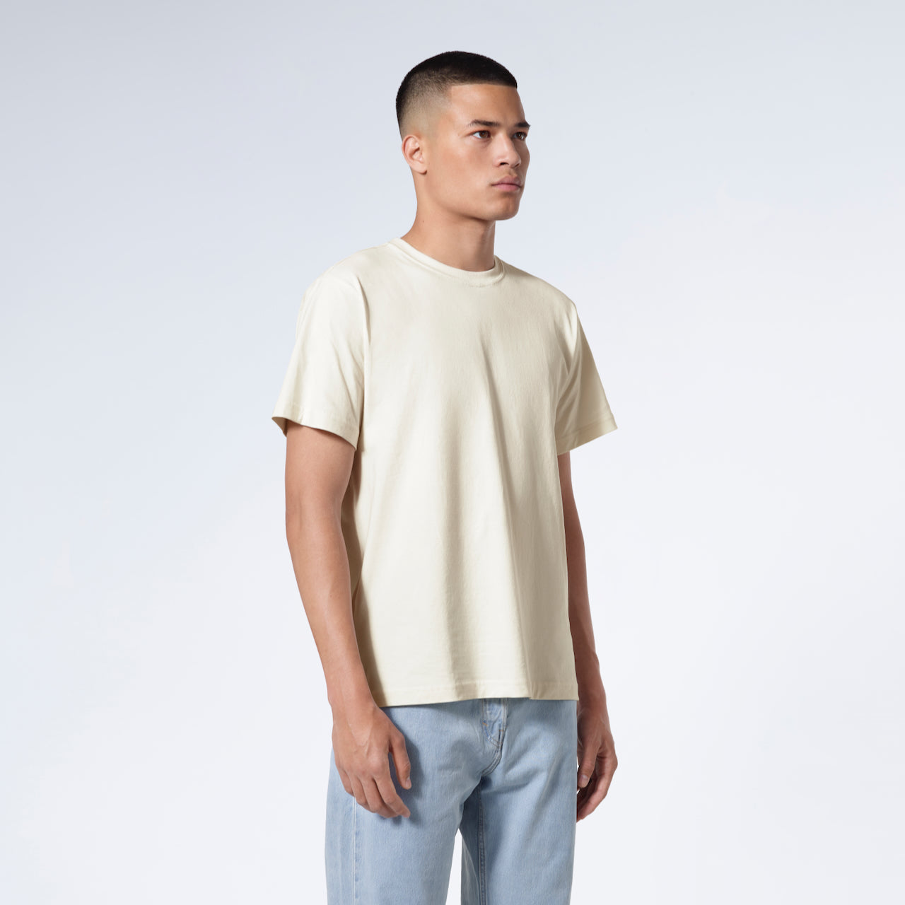 A man at an angle with his arms by his sides, modelling a plain ecru short sleeve organic cotton t shirt.