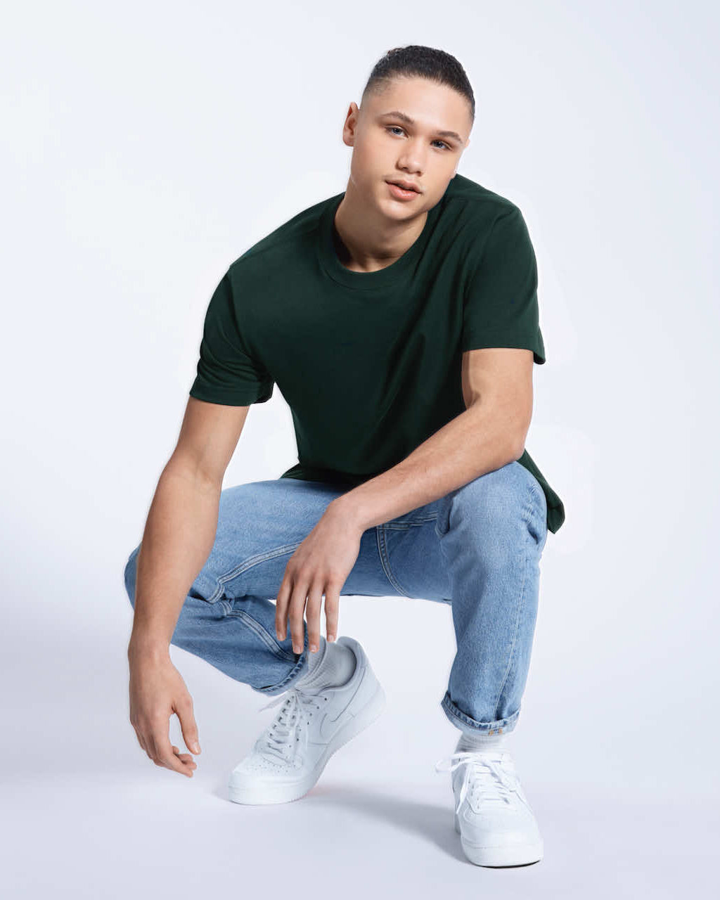 A man crouching down looking into the camera, modelling a plain green short sleeve organic cotton t shirt.