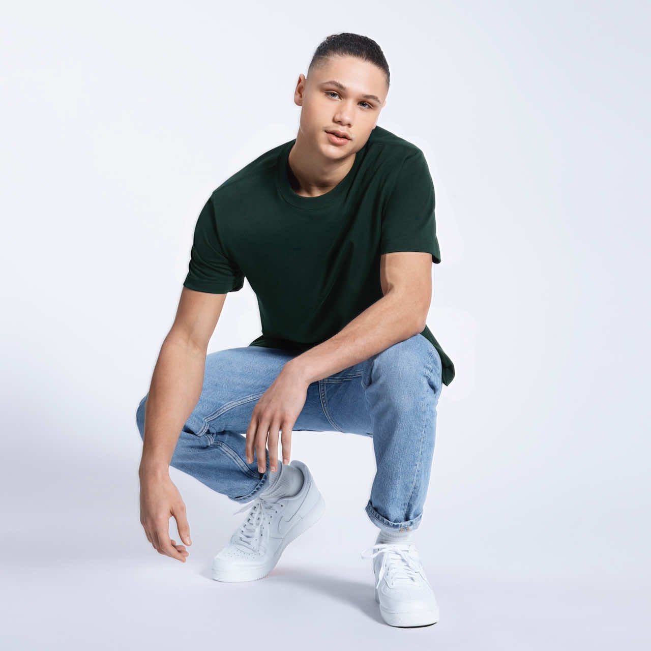 A man crouching down looking into the camera, modelling a plain green short sleeve organic cotton t shirt.