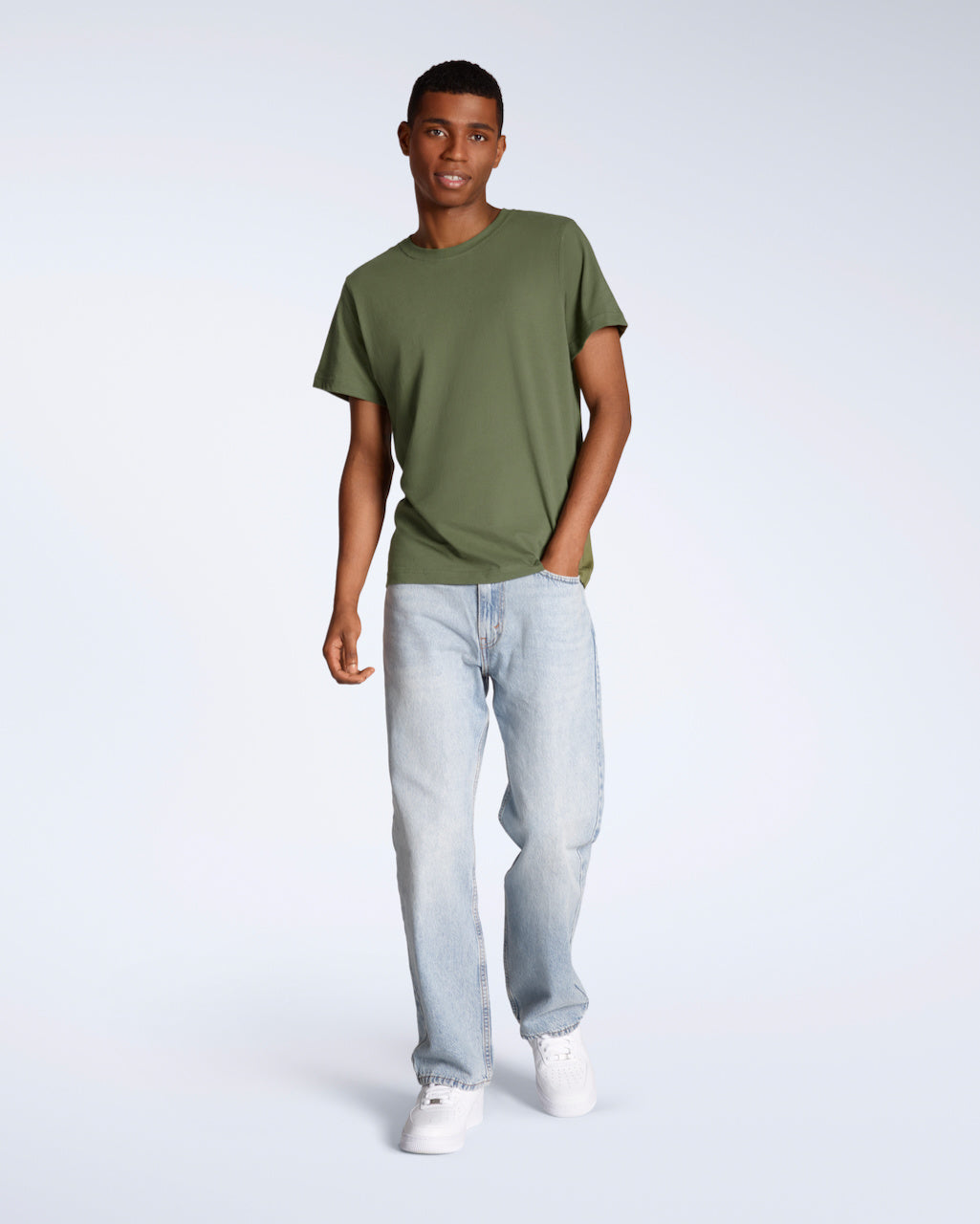 A full body photo of a man looking into the camera with his hand in his pocket, modelling a plain khaki green, short sleeve organic cotton t shirt with a pair of jeans.